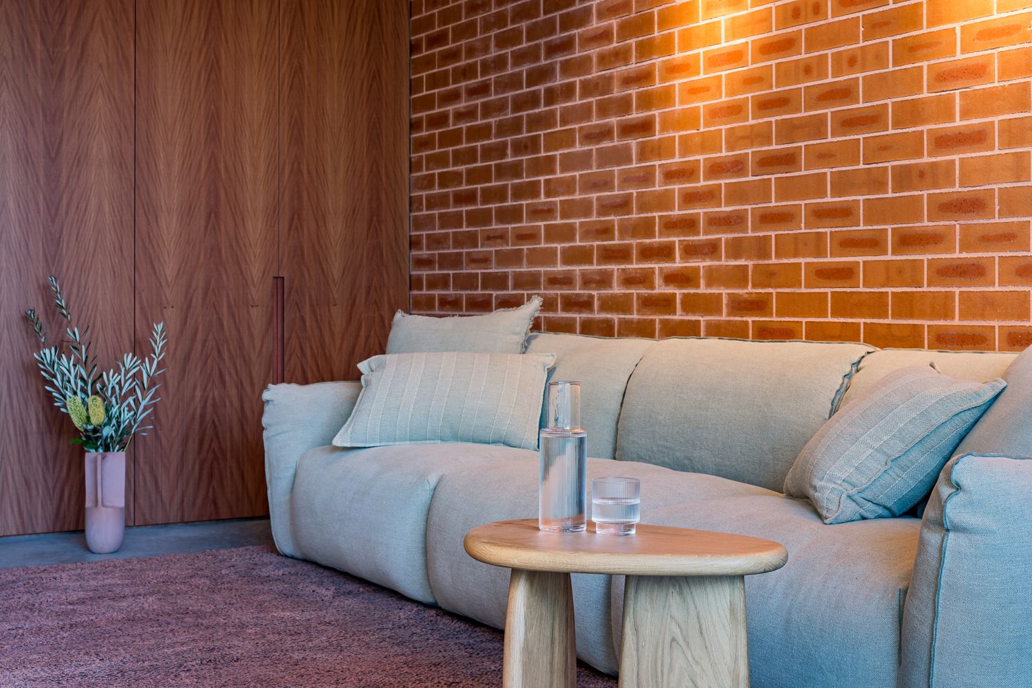 Soft sage green sofa with cushions against an orange brick wall and timber panelling, with a wooden side table and fresh flowers.