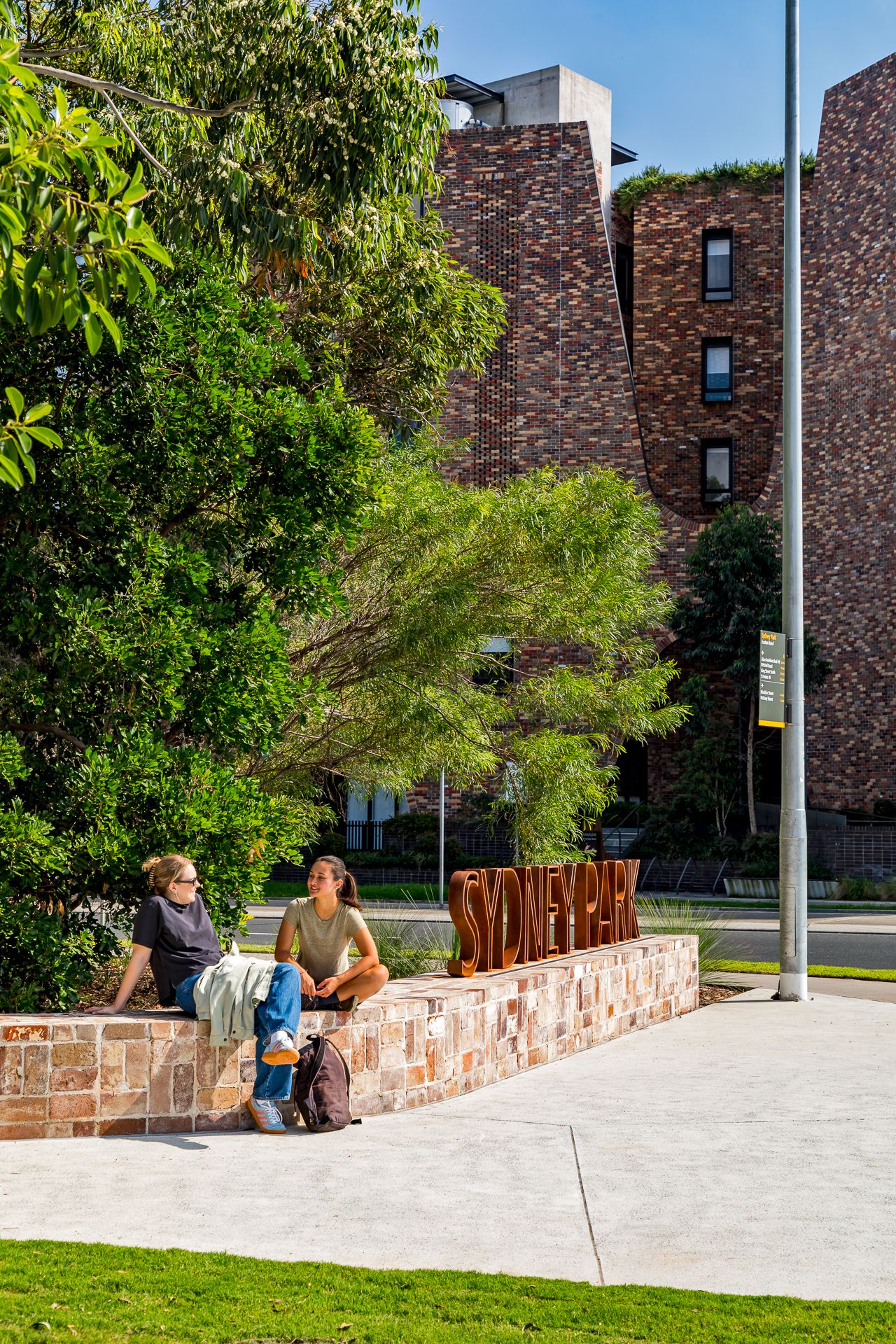 Two people relax on a sandstone wall beside the Sydney Park sign, with green trees and brick apartments behind.
