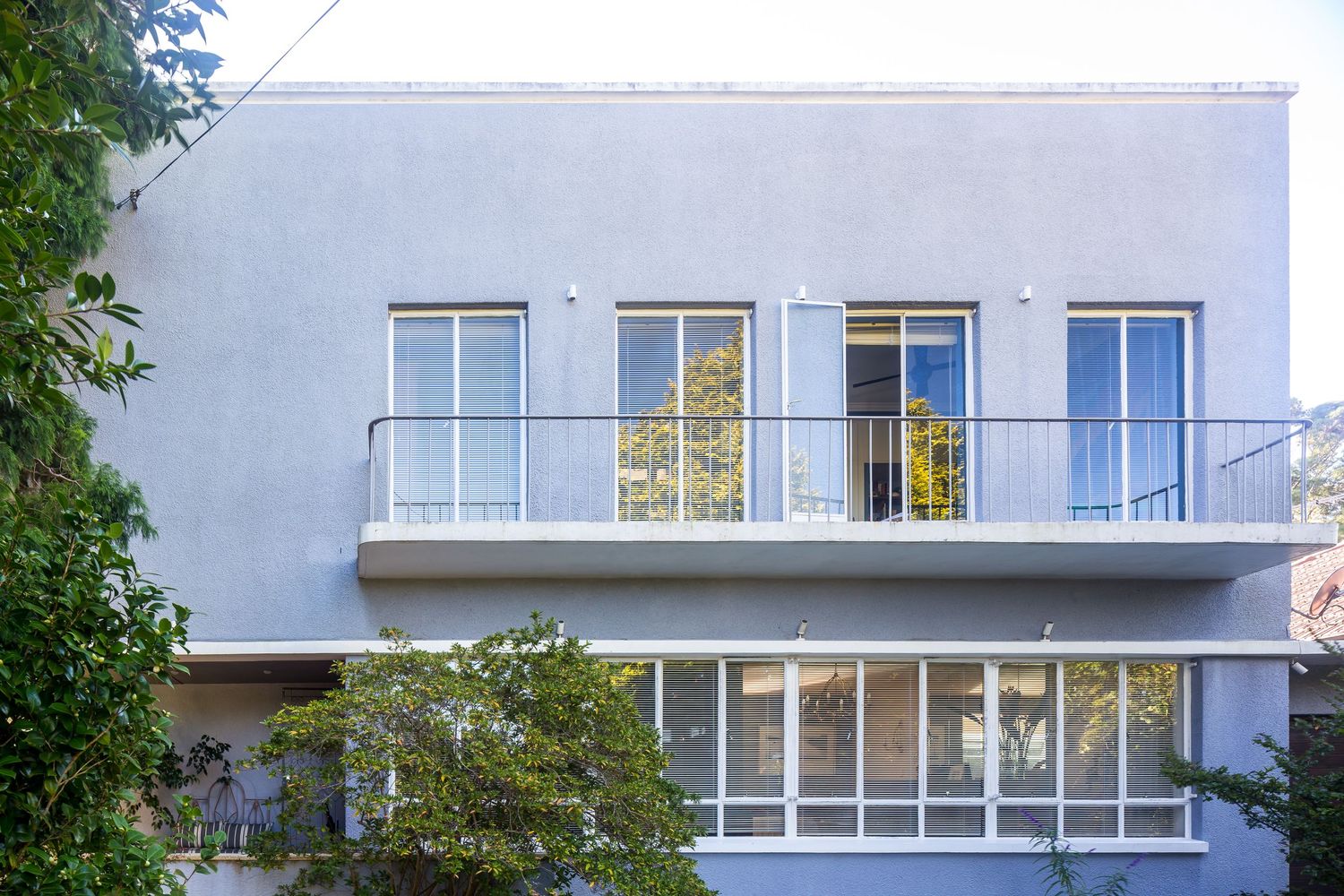 Light blue-gray modernist apartment building with a balcony, large rectangular windows, and green trees framing the facade.