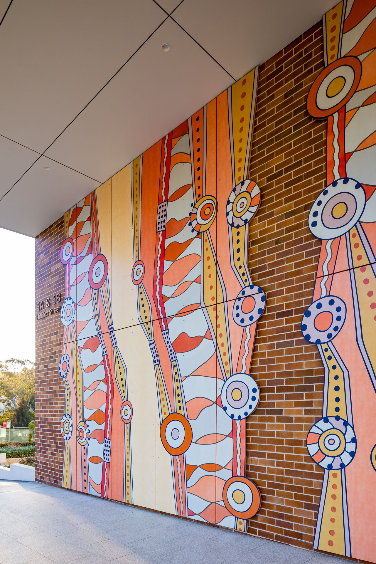 Colourful Aboriginal-style mural in orange, cream and blue dot patterns on a brick wall beneath a modern building overhang.