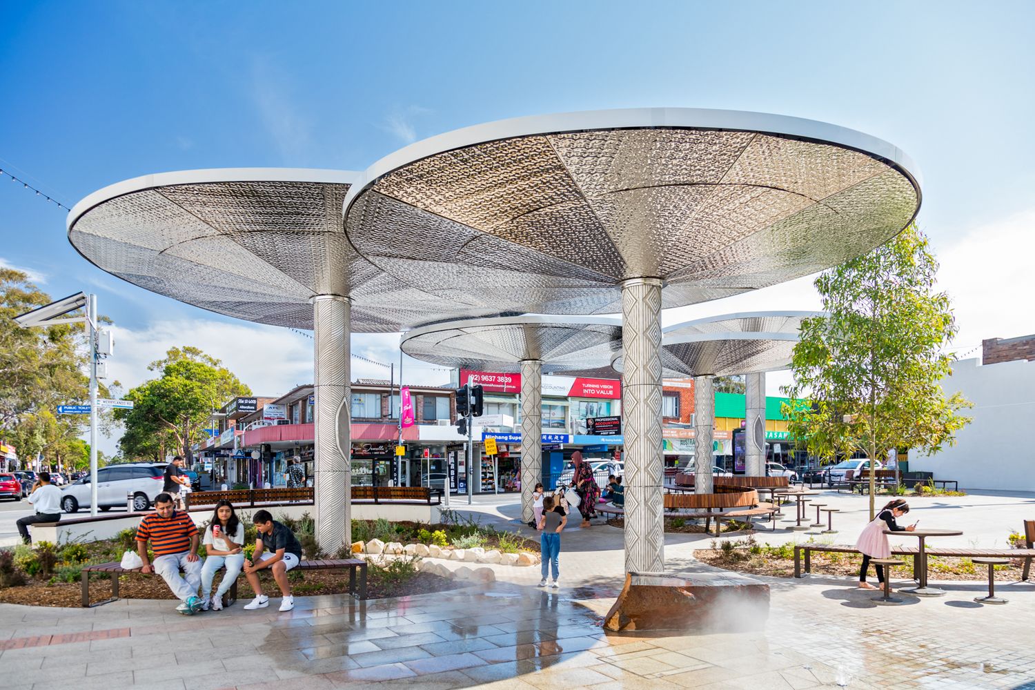 People relax on benches beneath large mushroom-shaped shade canopies in a sunny public plaza with shops in the background.