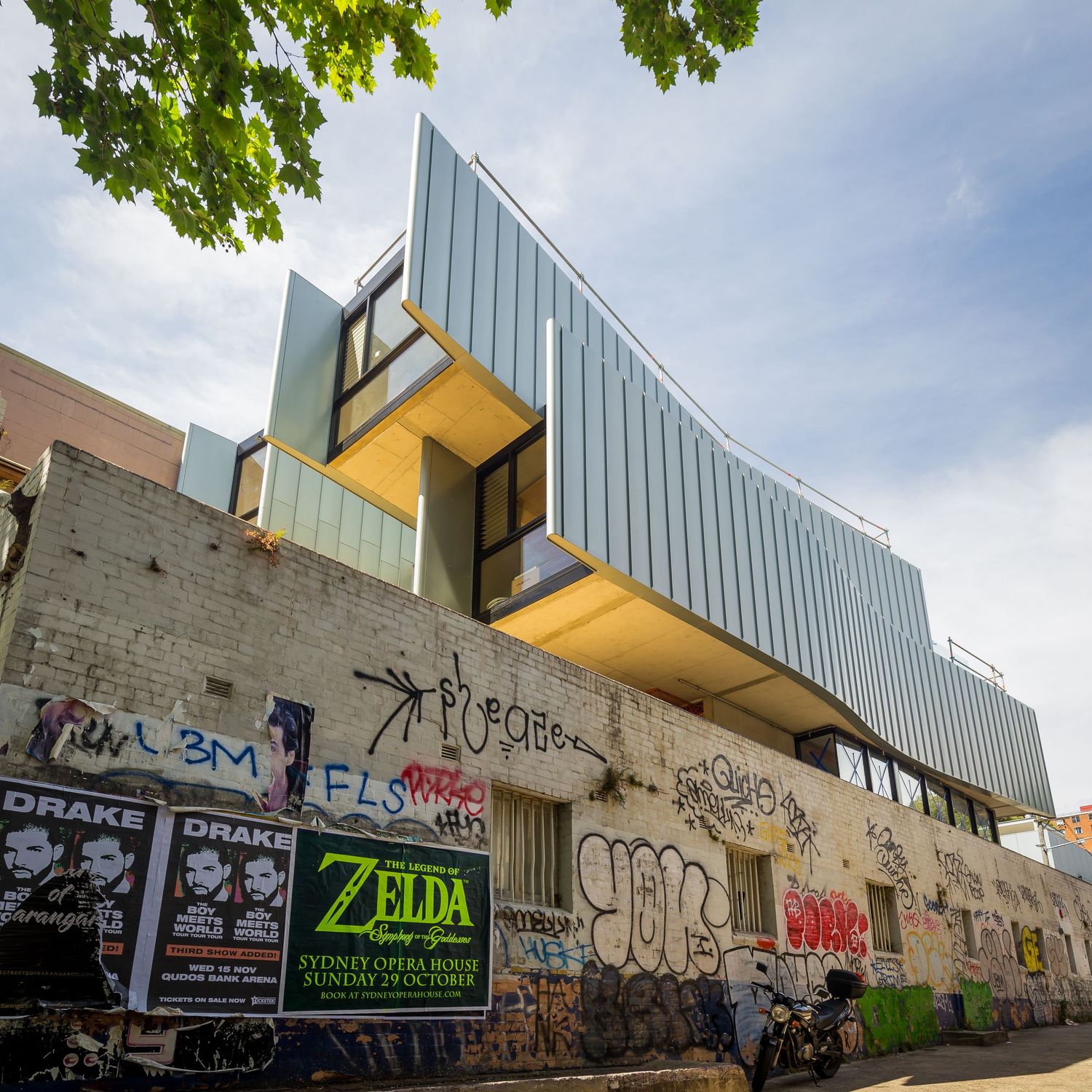 Modern corrugated metal building with cantilevered upper levels above a graffiti-covered brick wall with Zelda and band posters.