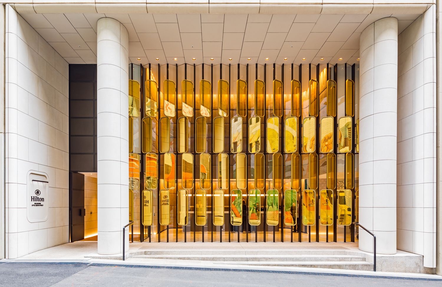 Upscale building entrance framed by tall white columns and a golden glass facade with vertical metal slats and greenery.