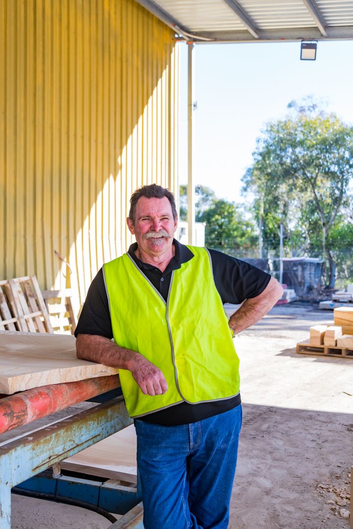 A smiling worker in a hi-vis yellow vest leans against a timber workbench outside a yellow shed on a sunny day.