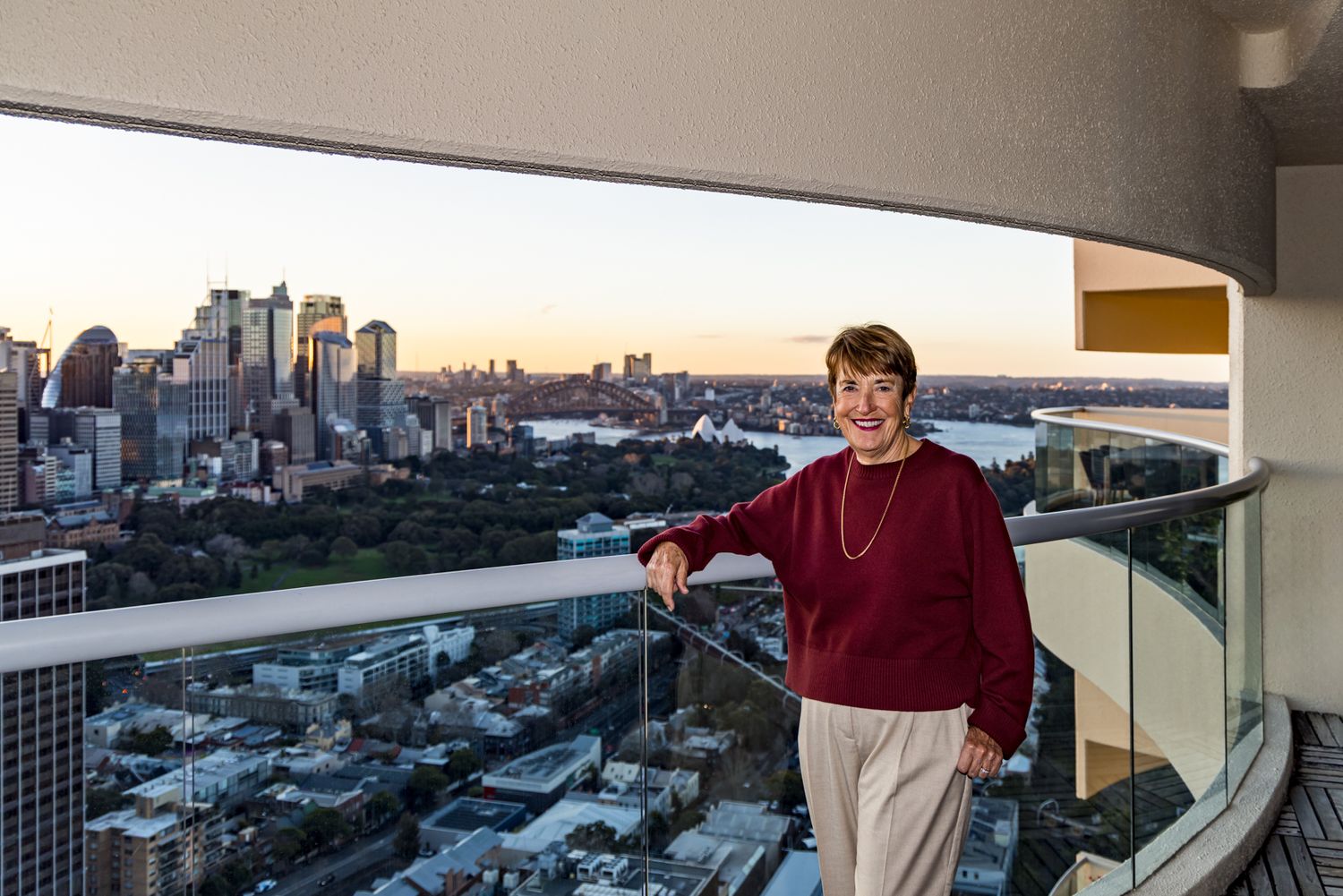 Smiling woman in a maroon sweater stands on a high-rise balcony overlooking a sweeping city skyline and harbour at dusk.