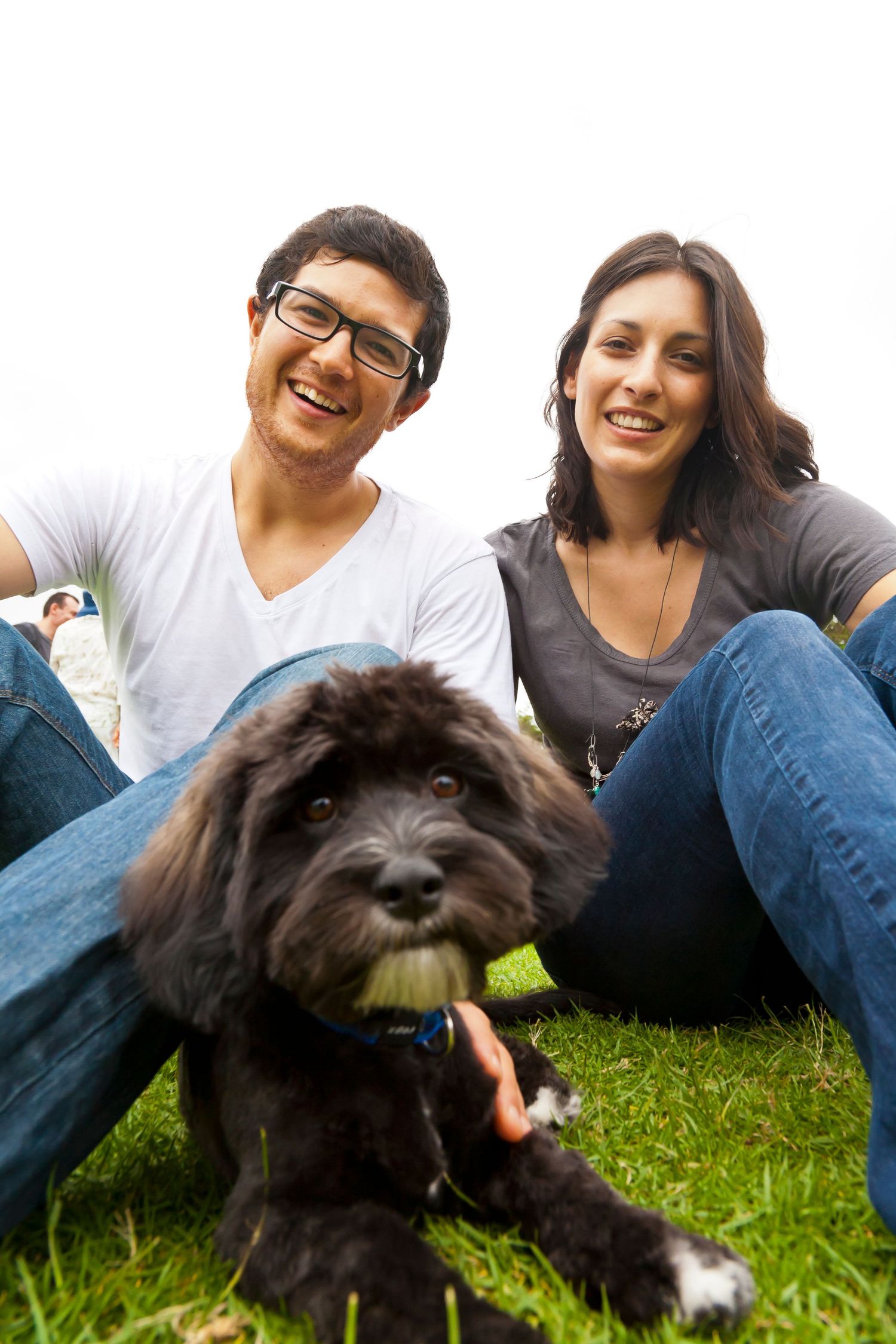 A smiling man with glasses and a smiling woman sit on the grass with a fluffy black dog in the foreground.