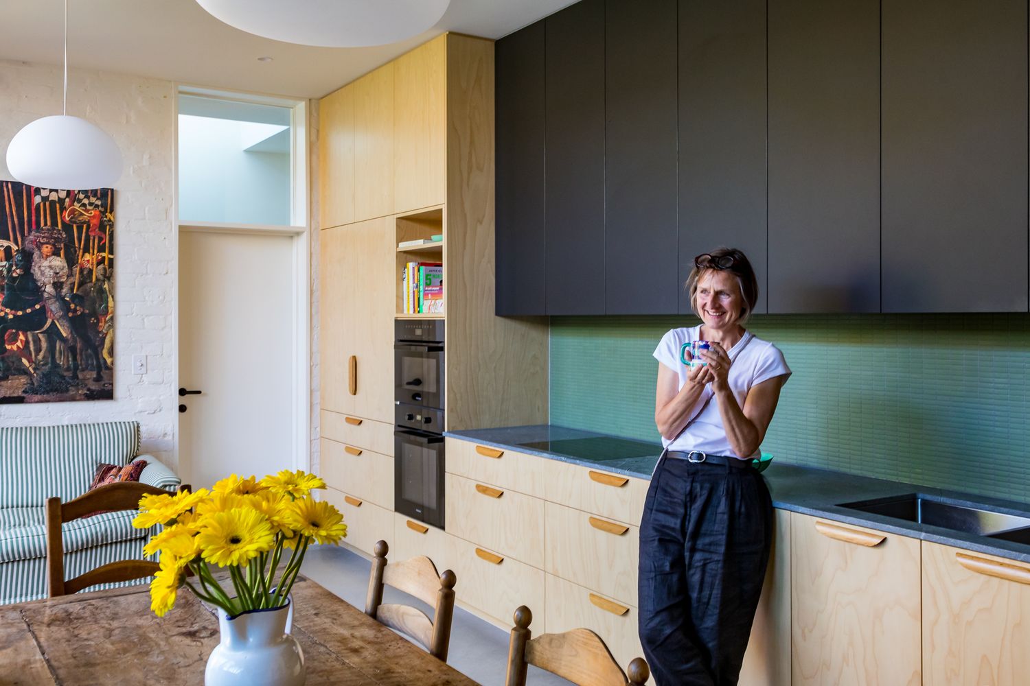 Smiling woman stands in a modern kitchen with timber cabinetry and a green splashback, near a table with a vase of yellow flowers.