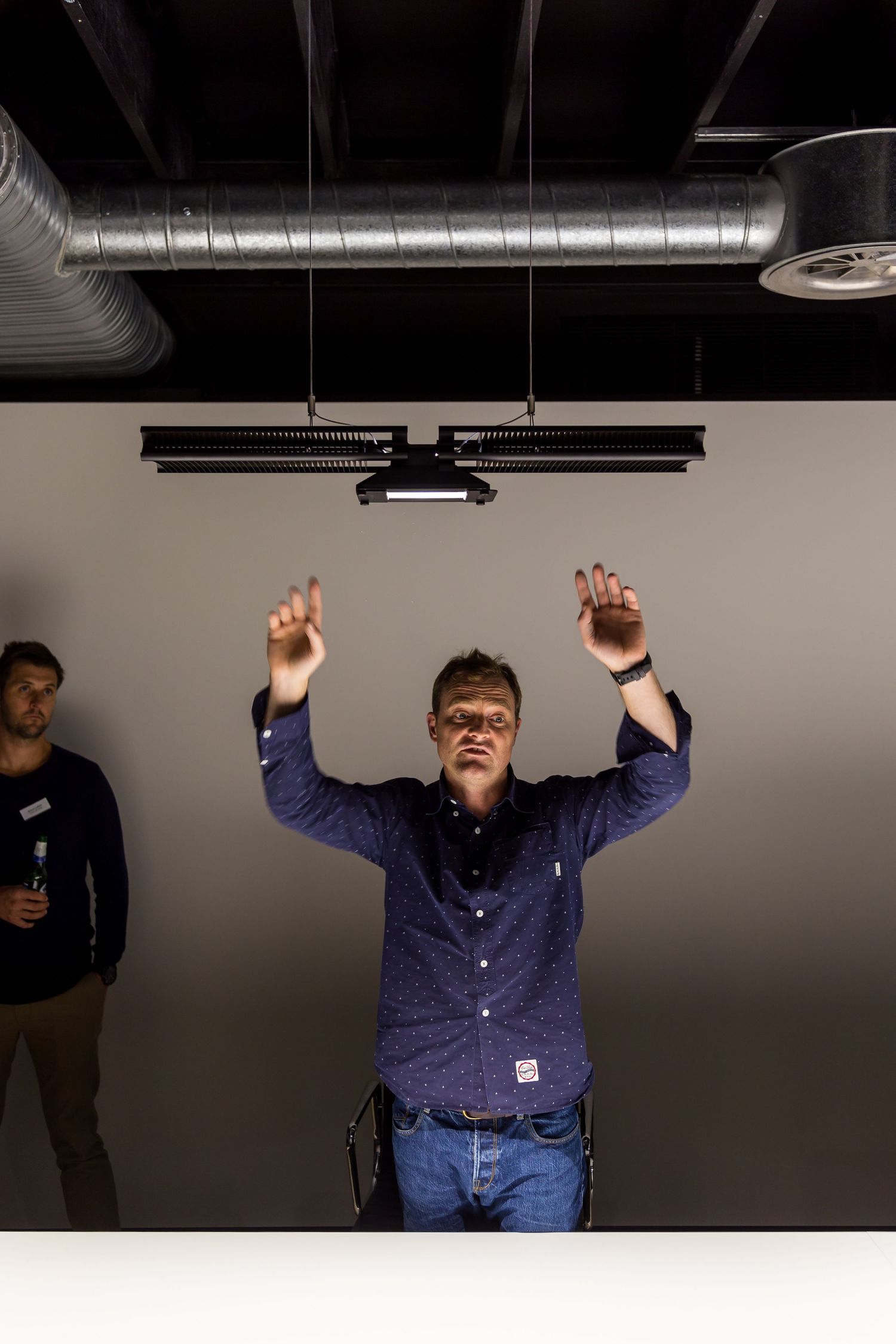 A man in a navy button-up shirt raises both hands above his head in a modern studio with exposed ductwork as a colleague stands behind him.