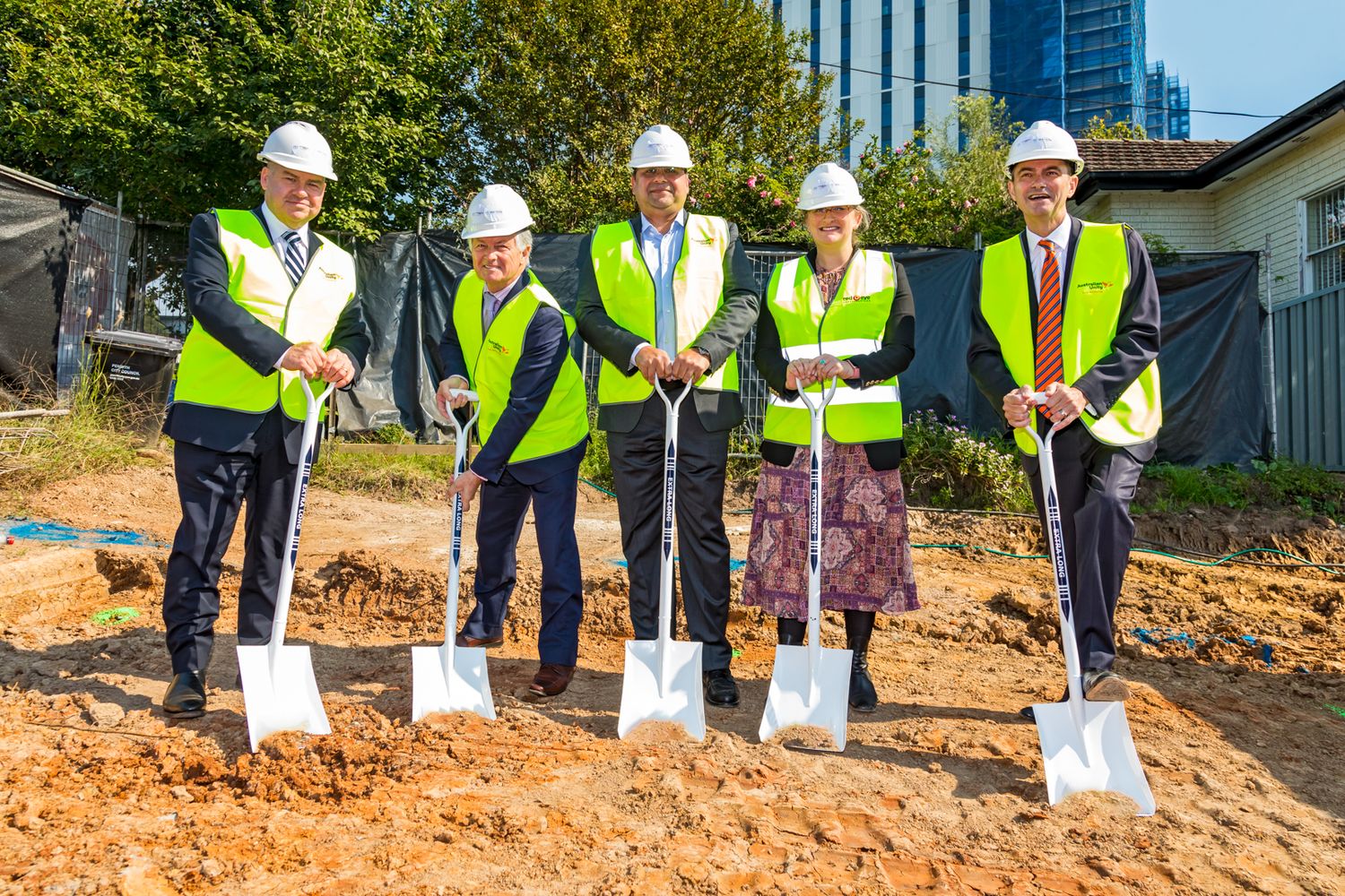 Five people in hard hats and yellow safety vests hold shovels at a groundbreaking ceremony on a construction site.