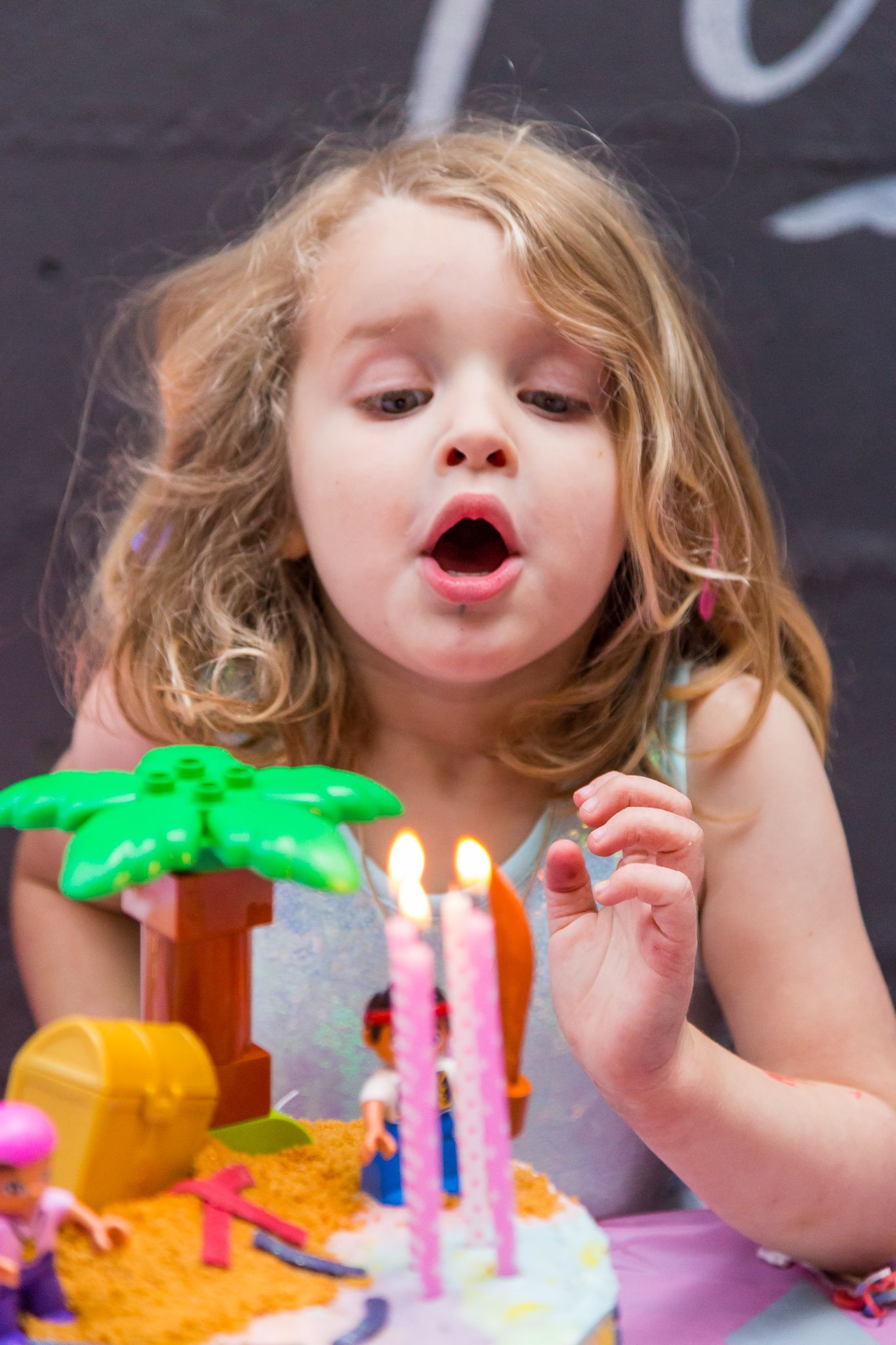 A young girl with wavy hair blows out two pink candles on a colorful birthday cake decorated with toy pirate figures and a treasure chest.