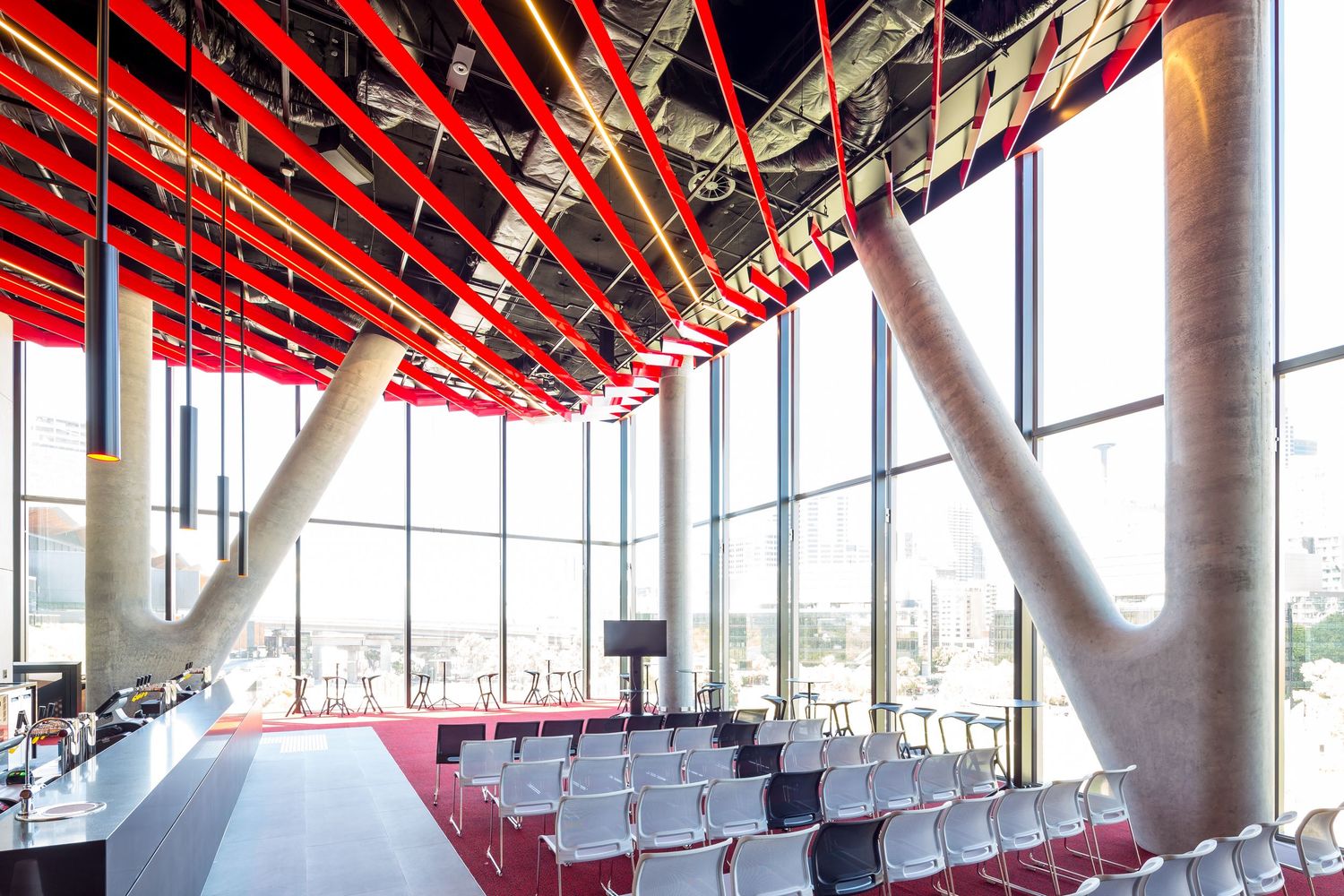 Conference hall with rows of white chairs, sloping concrete columns, floor-to-ceiling windows and a dramatic red slatted ceiling.