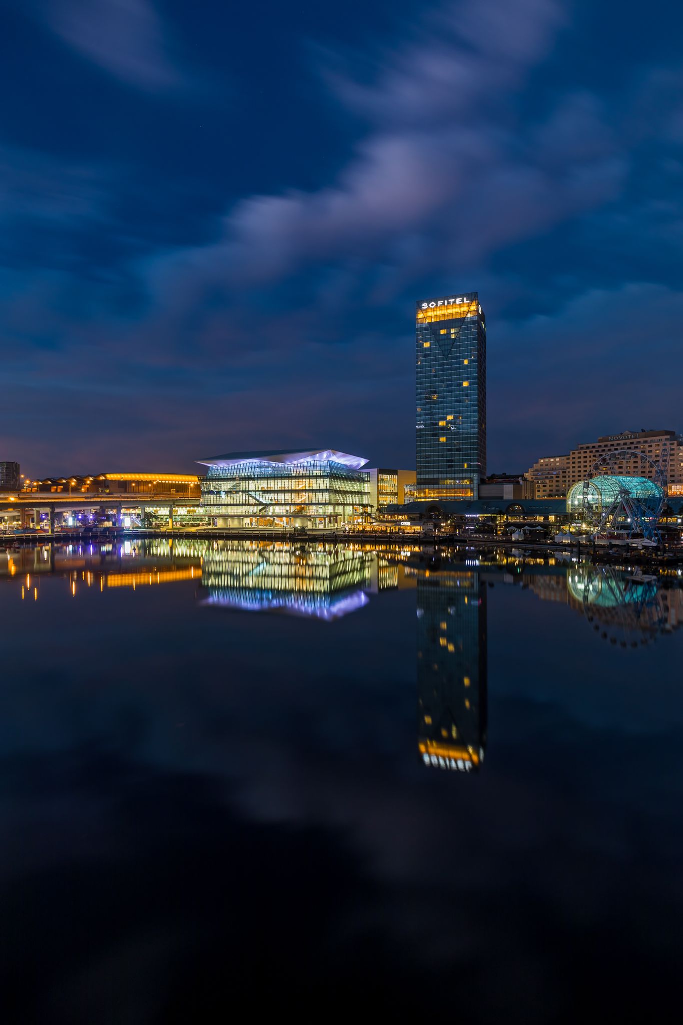 Harbourside skyline at twilight with an illuminated convention centre and tall hotel tower mirrored in dark, still water.