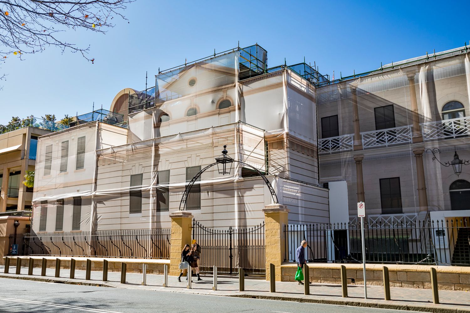 Sunlit historic building under restoration with scaffolding and sheeting, as pedestrians walk along the footpath past an ornate gate.