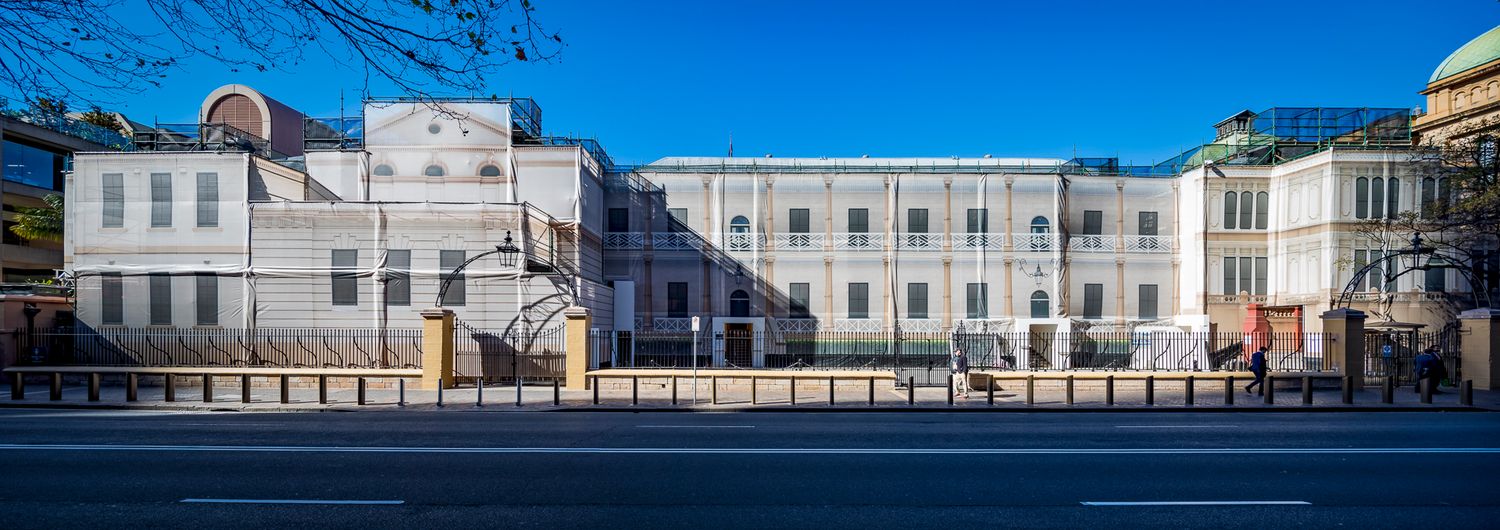 Panoramic view of a long heritage building and adjoining structures along a city street under a clear blue sky.