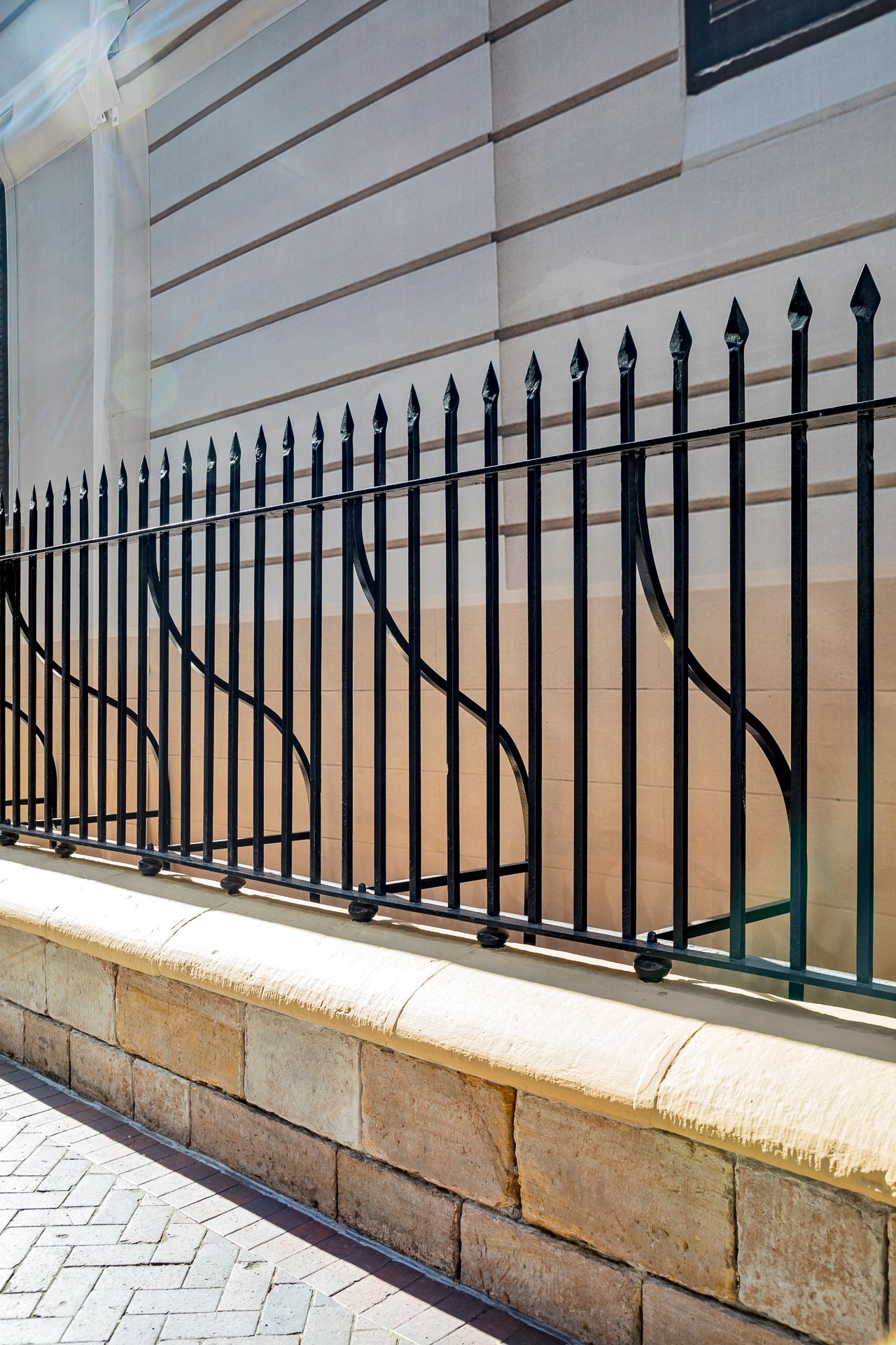 Black wrought iron fence with spear-tipped pickets and curved details atop a sandstone wall beside a paved footpath.