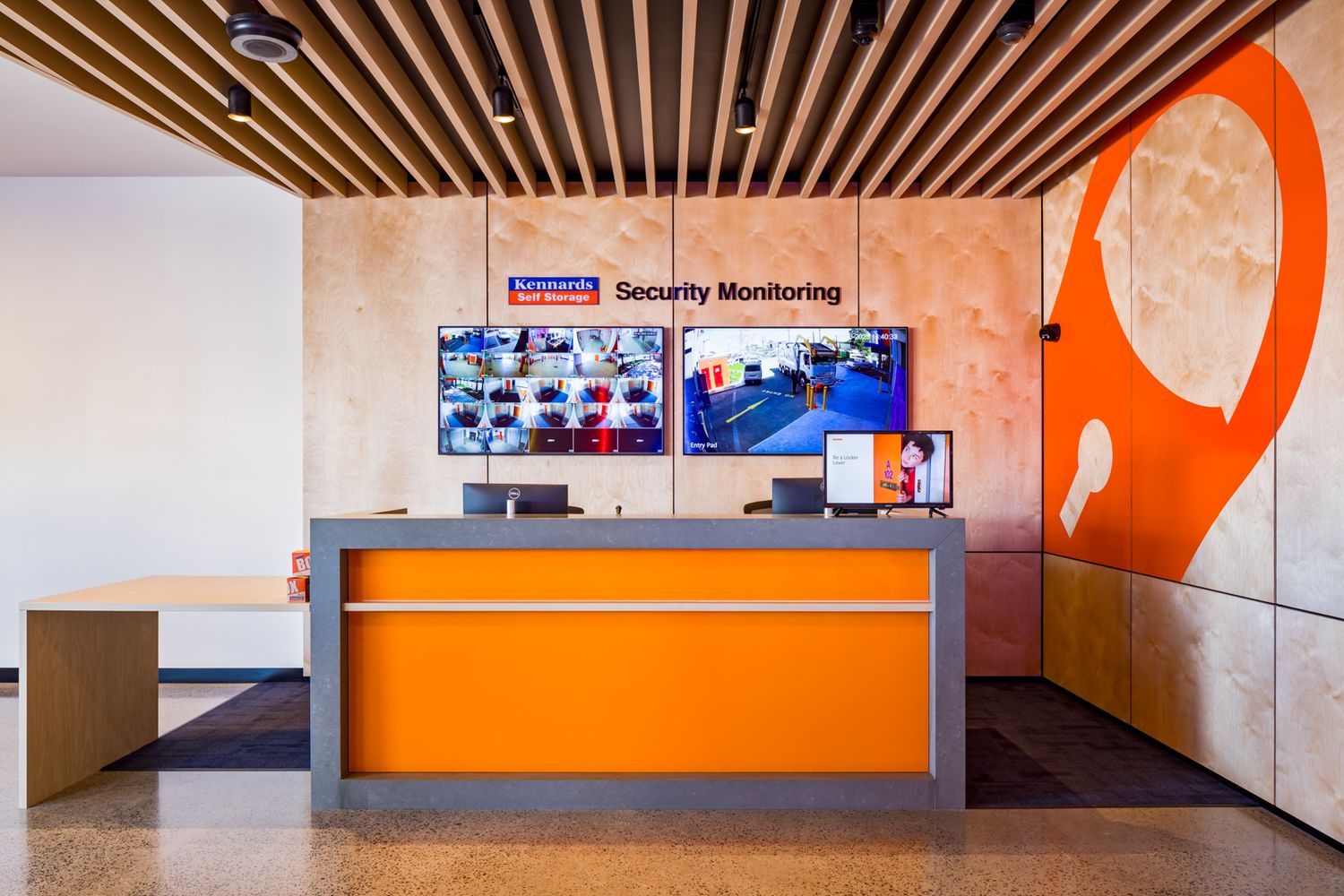 Orange reception desk beneath a Security Monitoring display of camera feeds in a modern self storage facility.