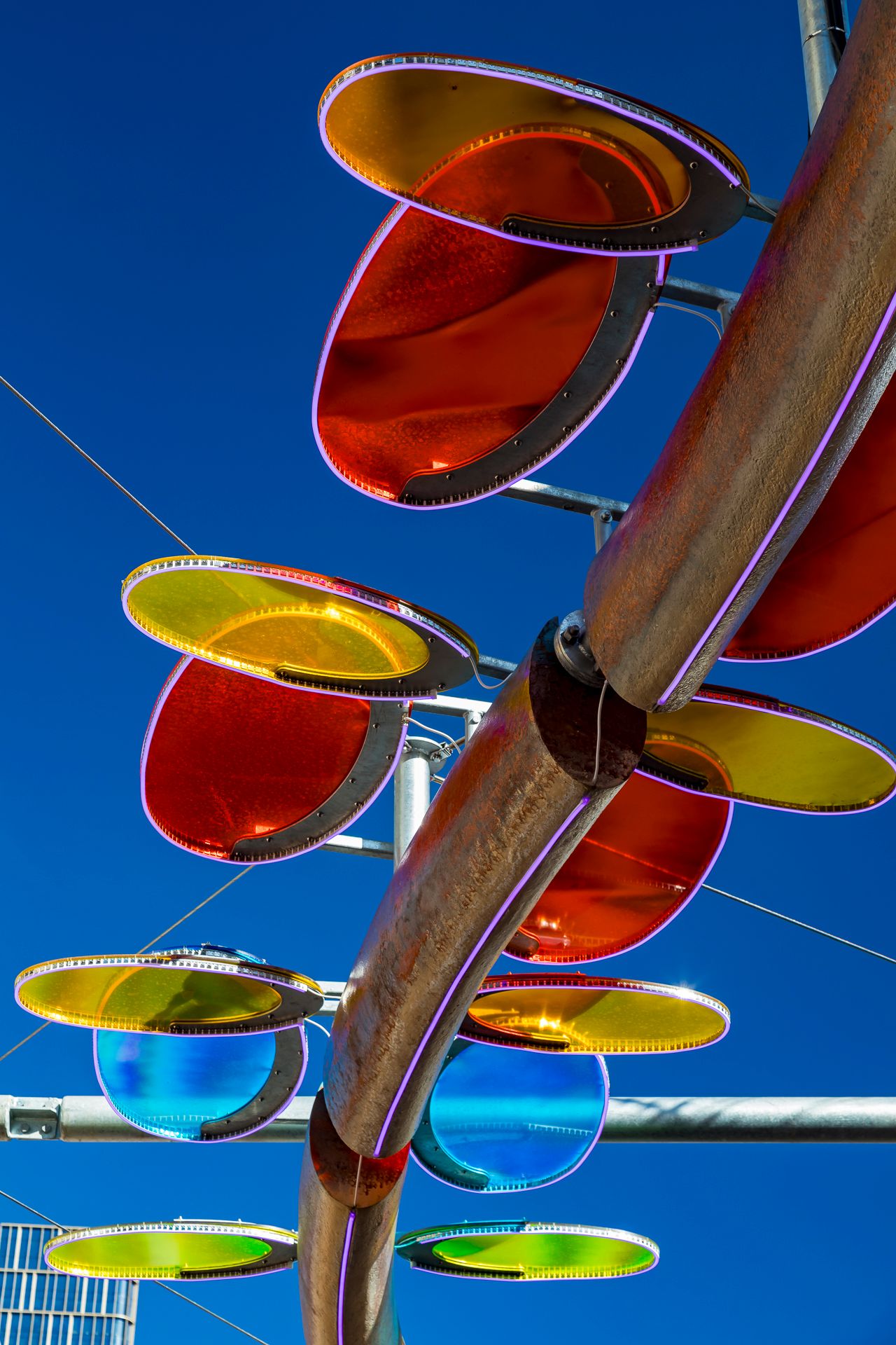 Red, orange, yellow, green and blue glowing discs attached to a copper-toned curved sculpture against a bright blue sky.