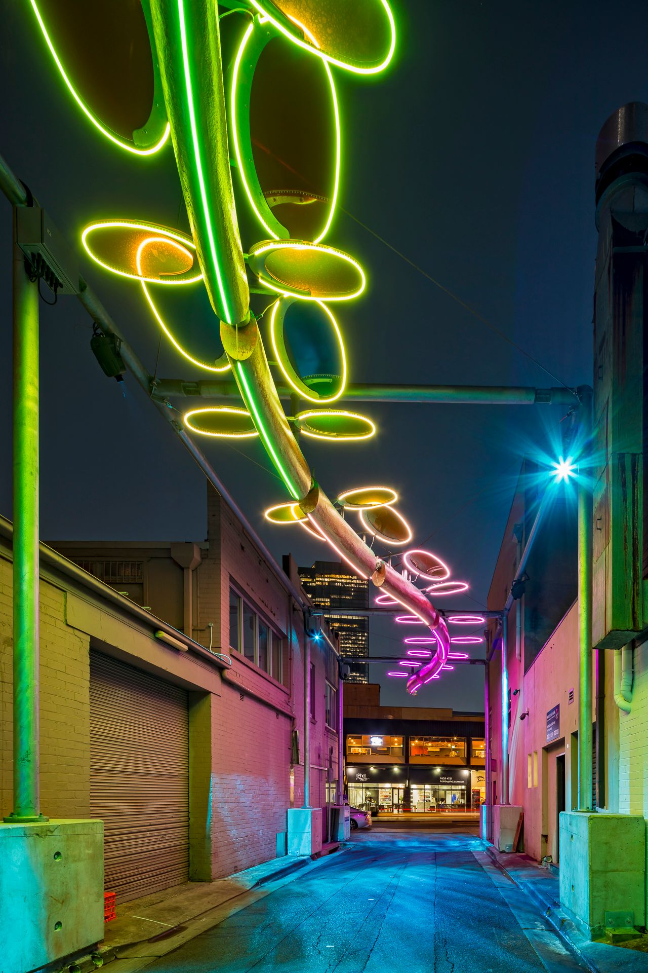 Close view of green and yellow neon oval rings on a suspended light sculpture over a city alley at night, with pink glow beyond.