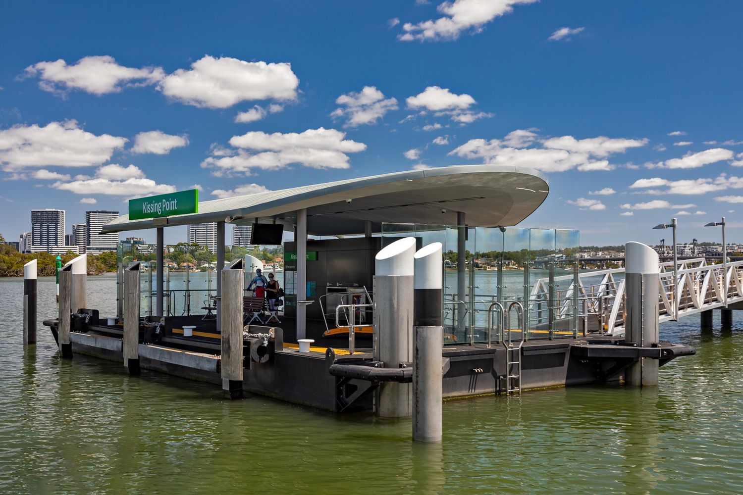 Kissing Point ferry wharf with a sweeping canopy and mooring piles floating on green river water beneath a blue sky with clouds.