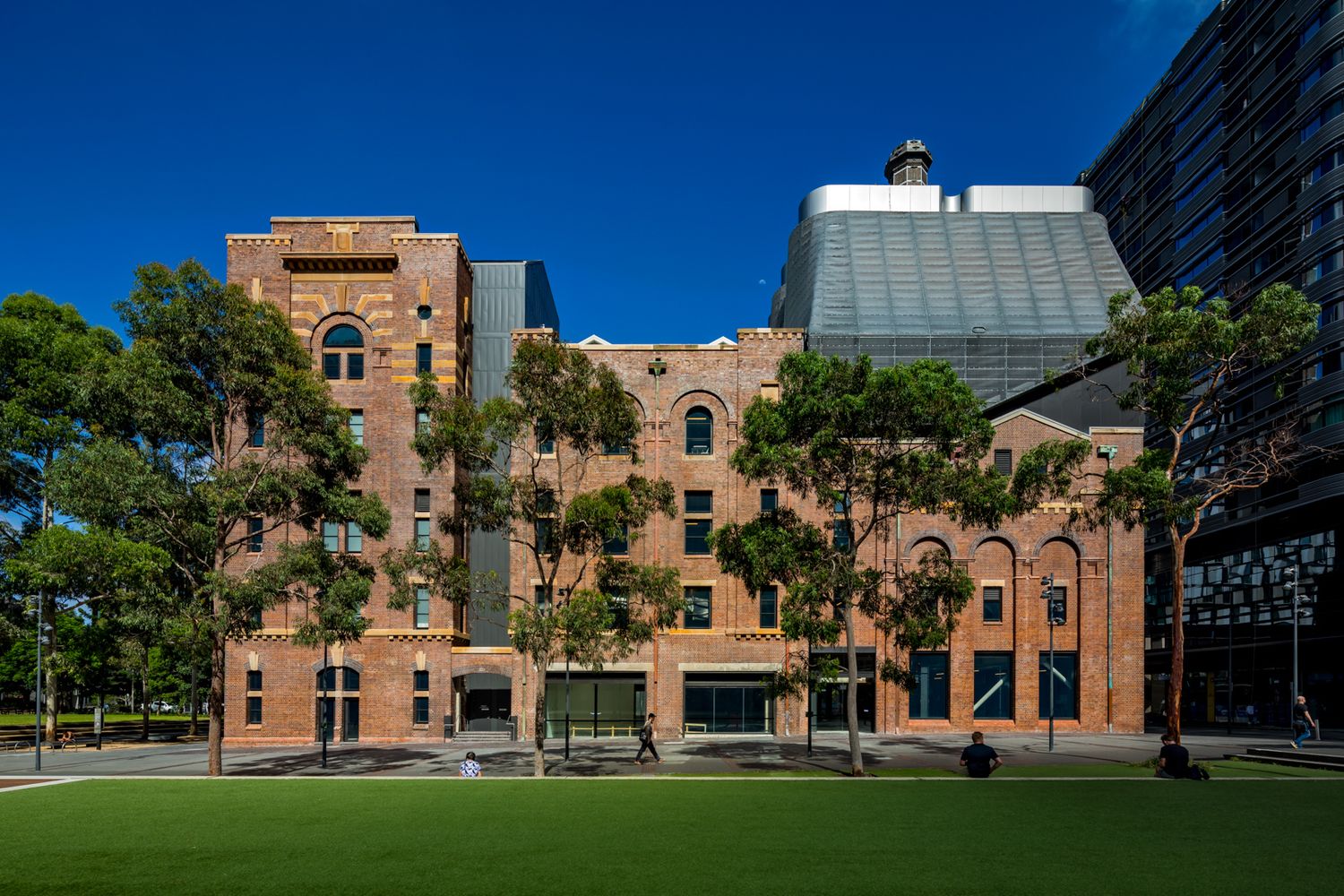 Restored red brick heritage warehouse with arched windows beside tall trees and a green lawn, next to modern glass buildings.