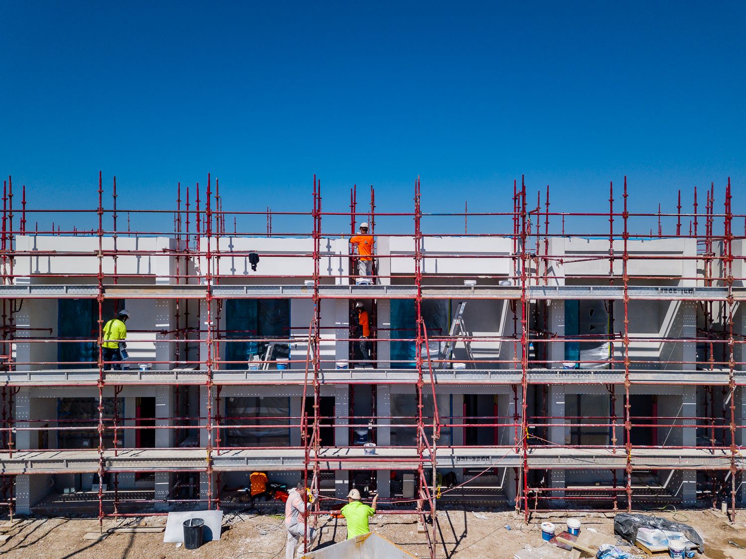 Construction workers in high-visibility vests on red scaffolding surrounding a multi-level townhouse building under a clear blue sky.