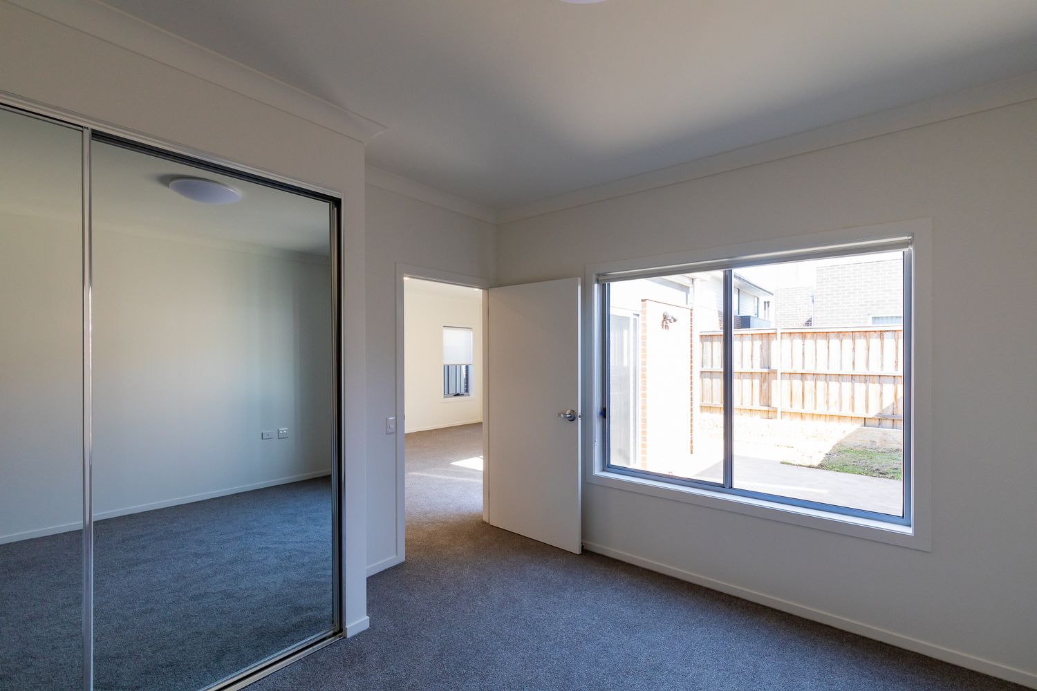 Bright empty bedroom with grey carpet, mirrored wardrobe doors and a large window overlooking a fenced backyard.