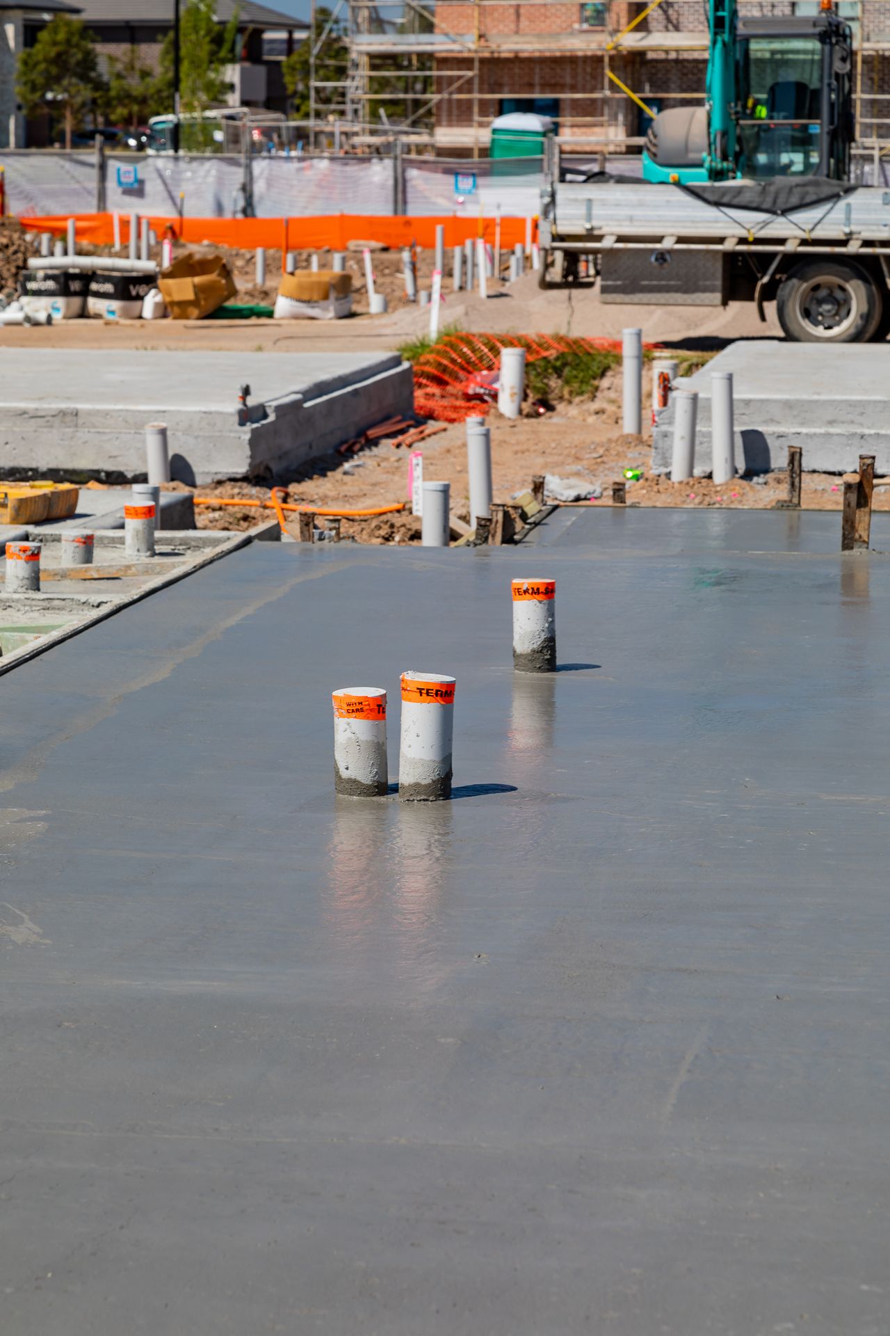 Smooth wet concrete slab with orange-banded pipes in the foreground and construction machinery working in the background.