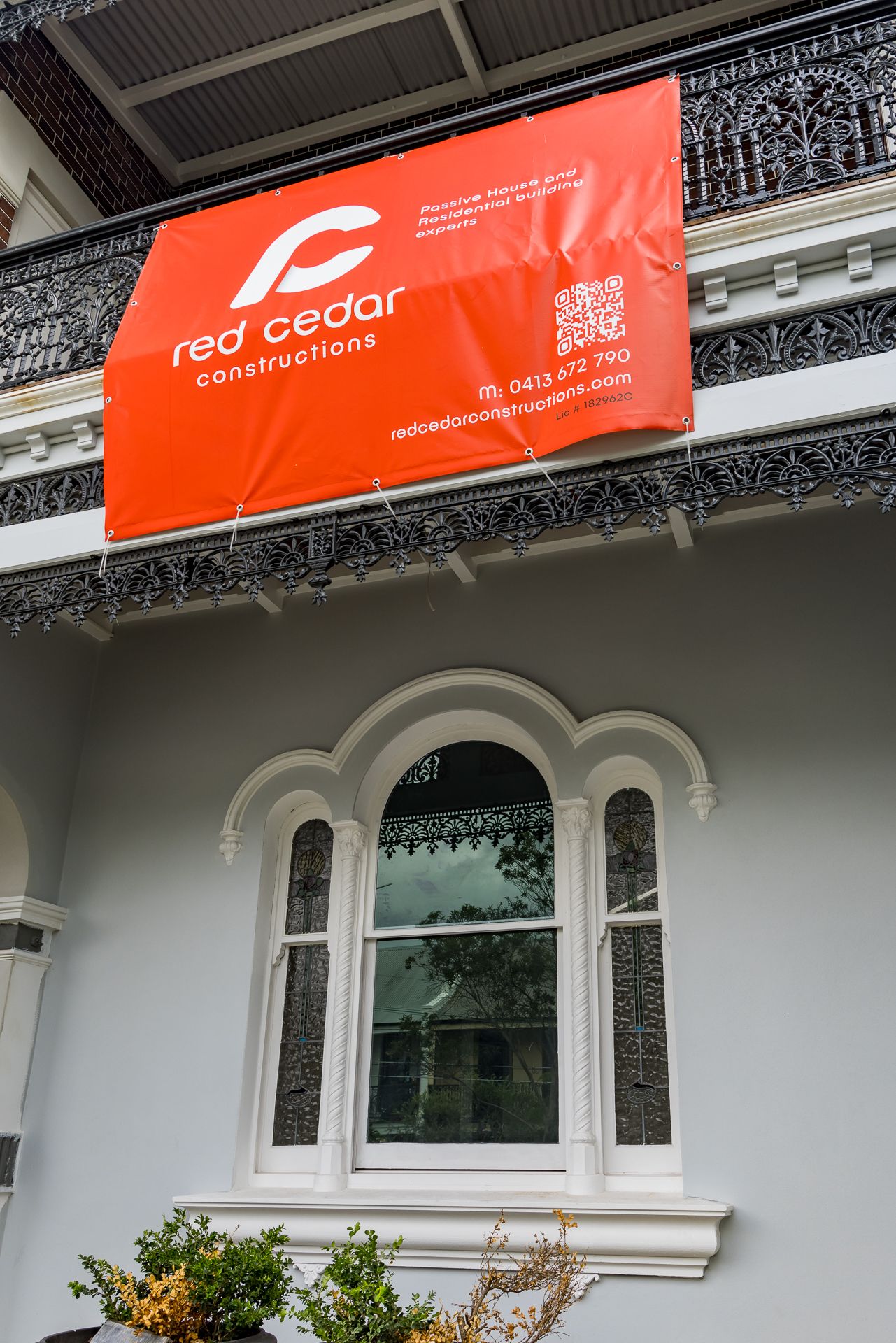 Red Cedar Constructions banner hung from an ornate cast iron balcony above a heritage arched window with leadlight side panels.