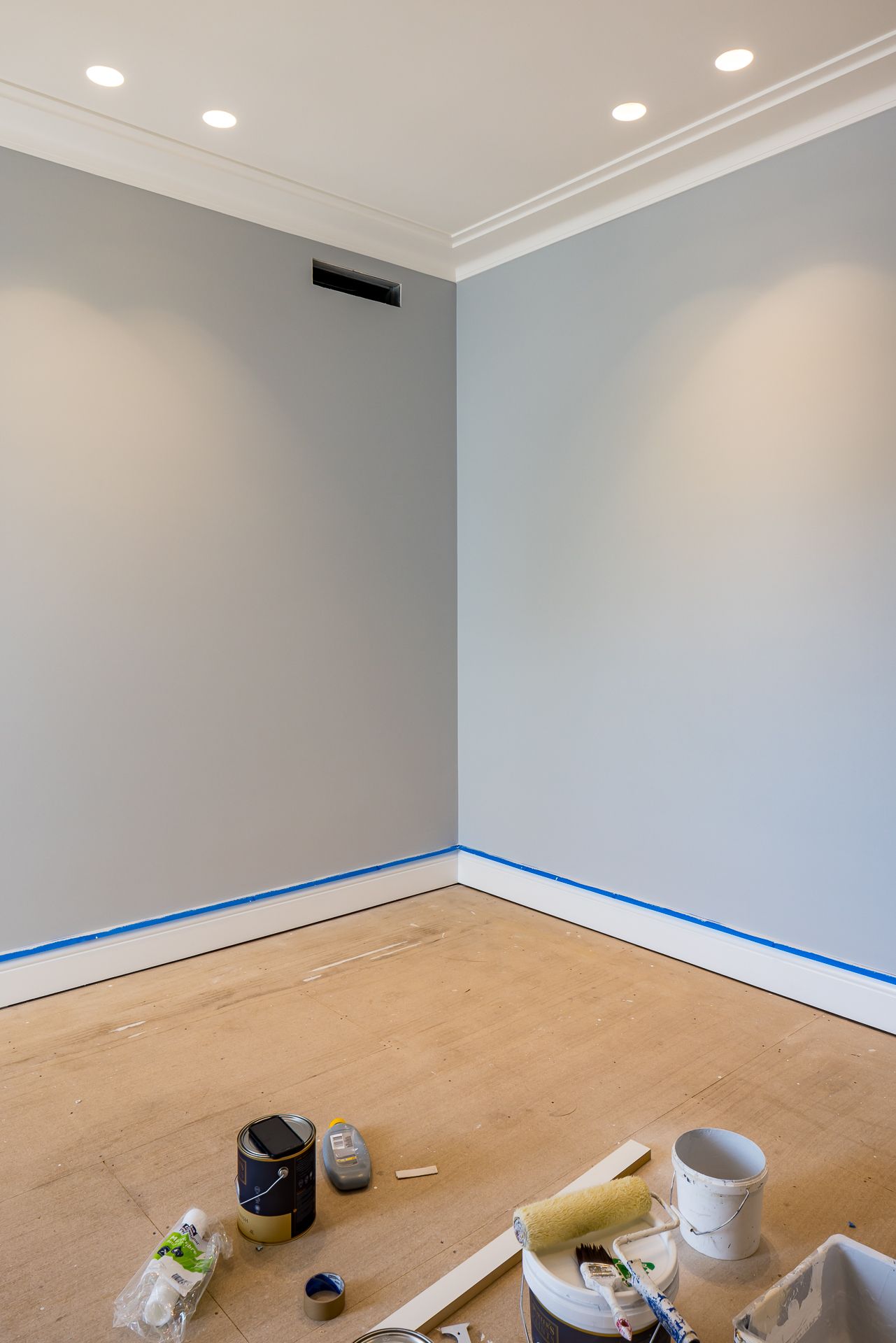 Freshly painted grey room with white cornice, recessed ceiling lights, blue masking tape and painting supplies on a bare timber floor.