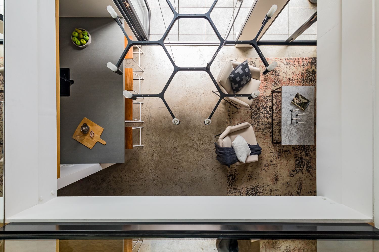 Overhead view of a modern living room with a hexagonal glass skylight, cushioned armchairs, a coffee table and a patterned area rug.