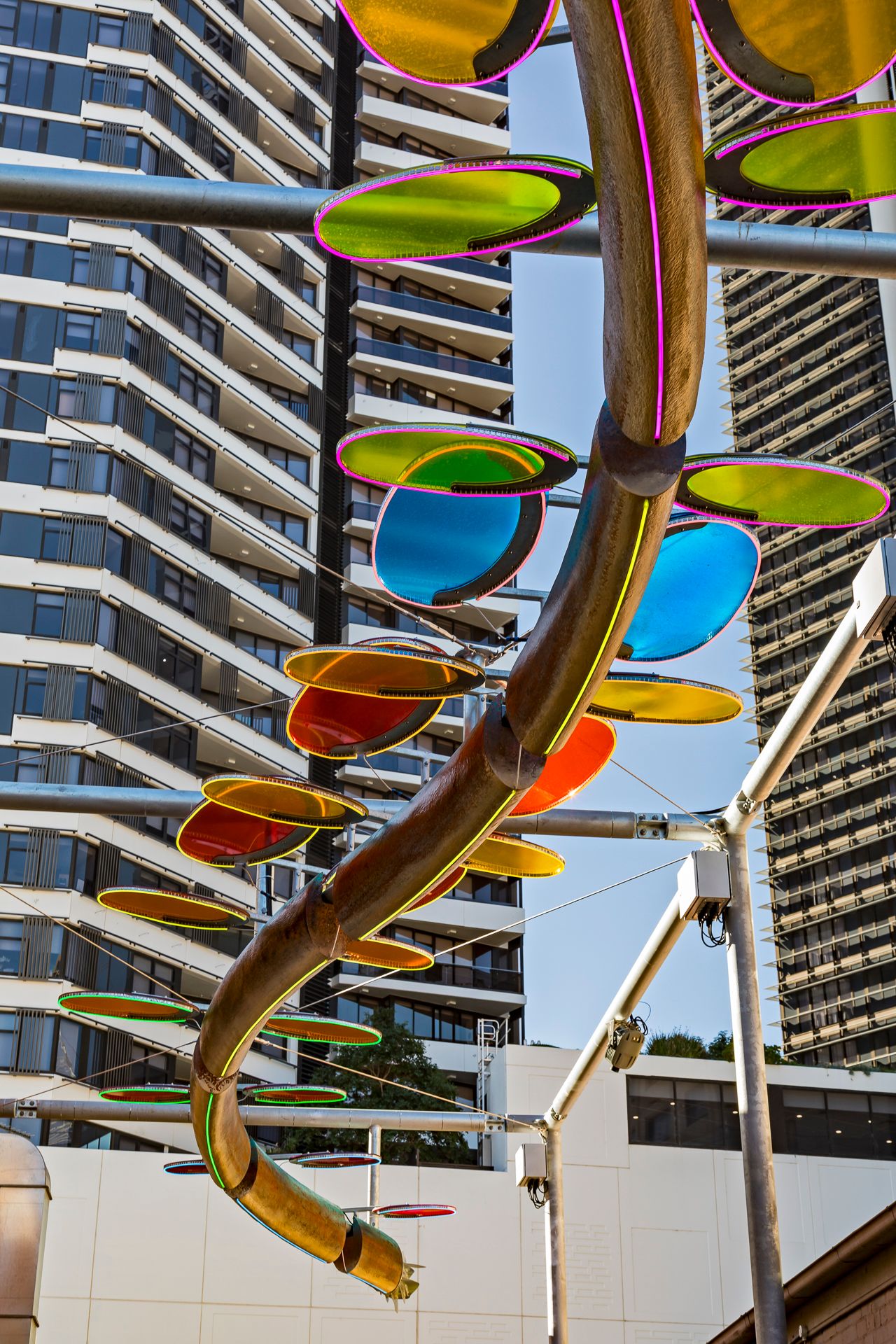 Spiral serpent sculpture with green, blue and orange oval discs suspended in front of tall high-rise apartment towers.