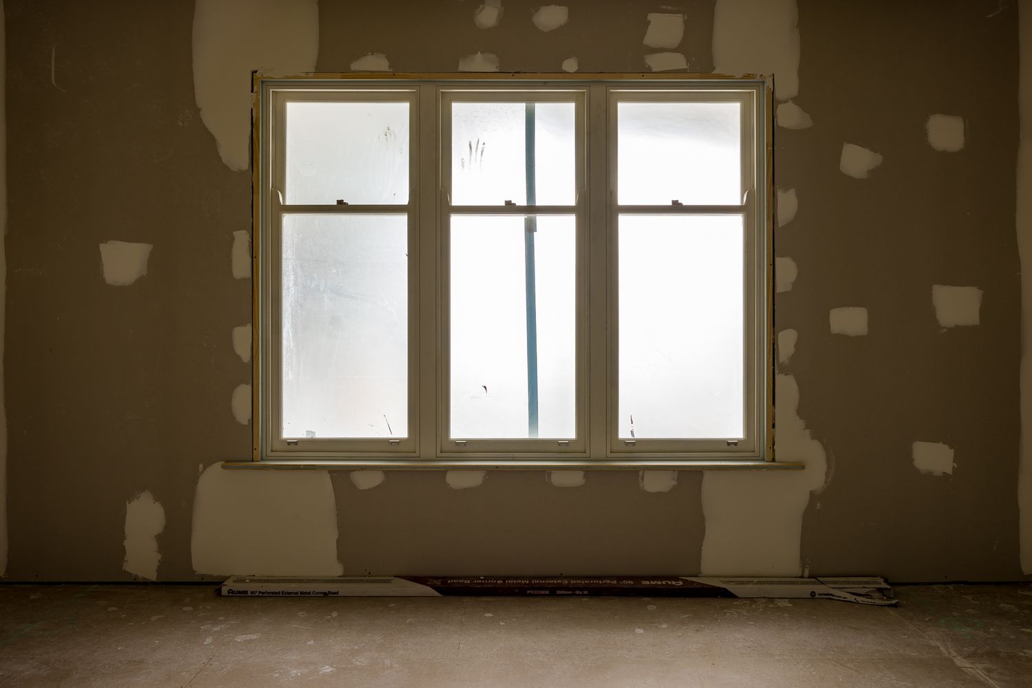 Large white-framed window with frosted glass in a gutted room with patched plaster walls and a bare concrete floor.