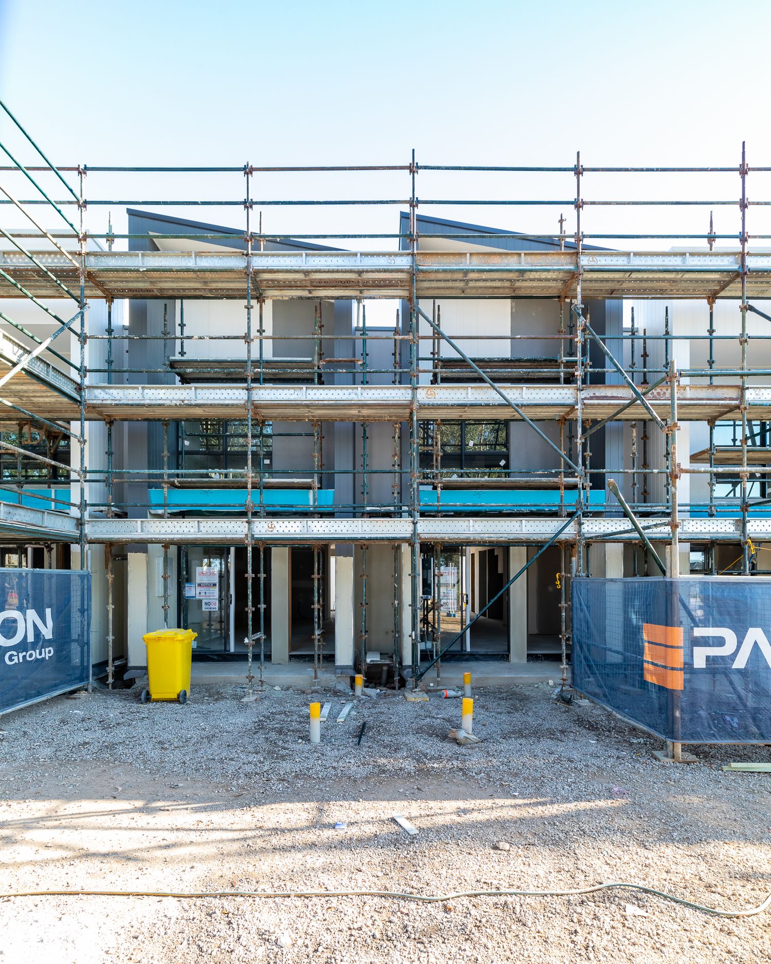 Multi-storey townhouse under construction with scaffolding, safety barriers and a yellow waste bin on a building site.