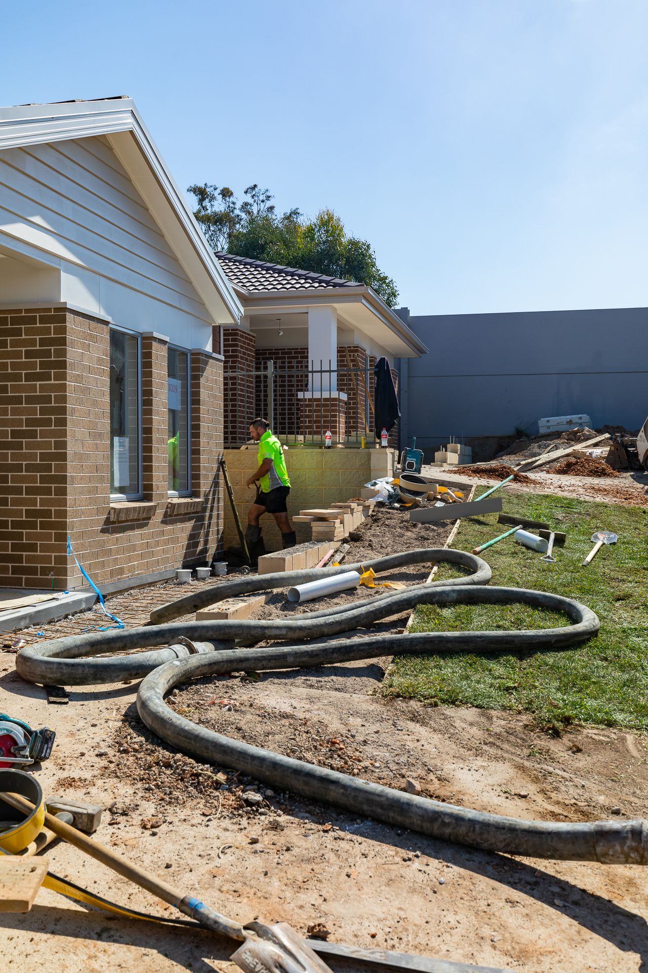 Construction worker in hi-vis shirt beside a new brick home with large black hoses laid across the dirt and grass.