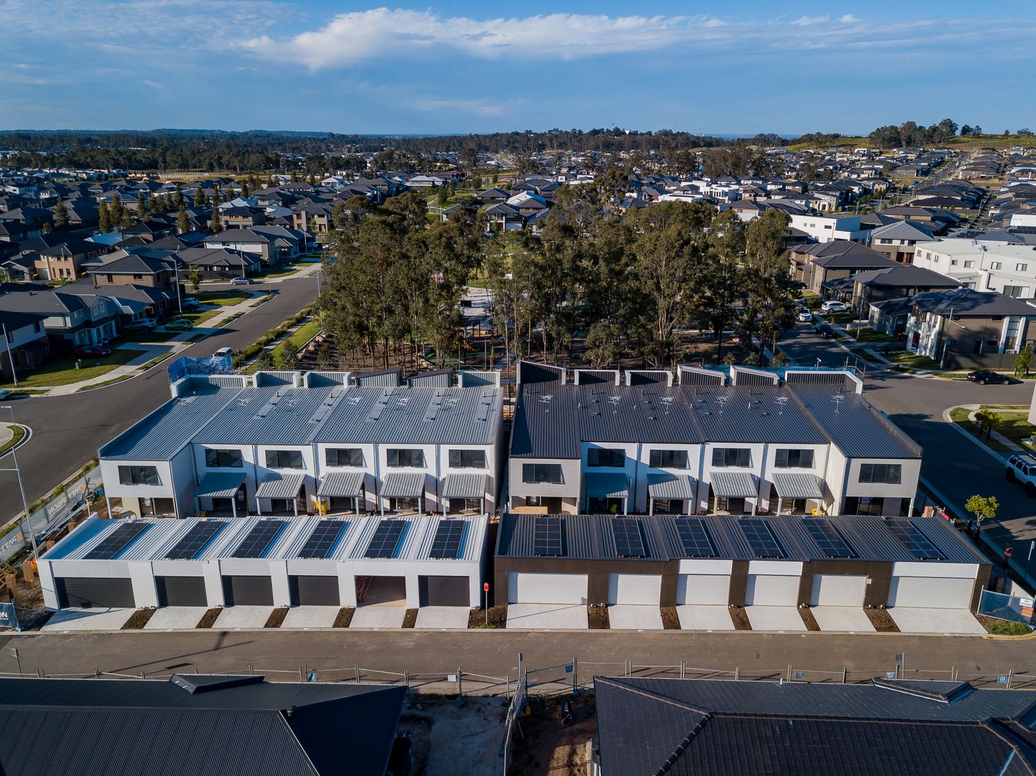 Aerial view of new modern townhouses with dark roofs and garages in a growing suburban housing development surrounded by trees.