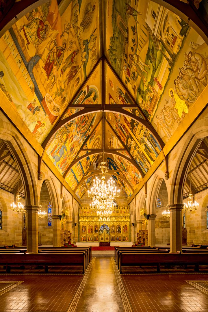 Church interior with a painted vaulted ceiling of colorful sacred murals, a glowing chandelier, wooden pews and an ornate golden altar.