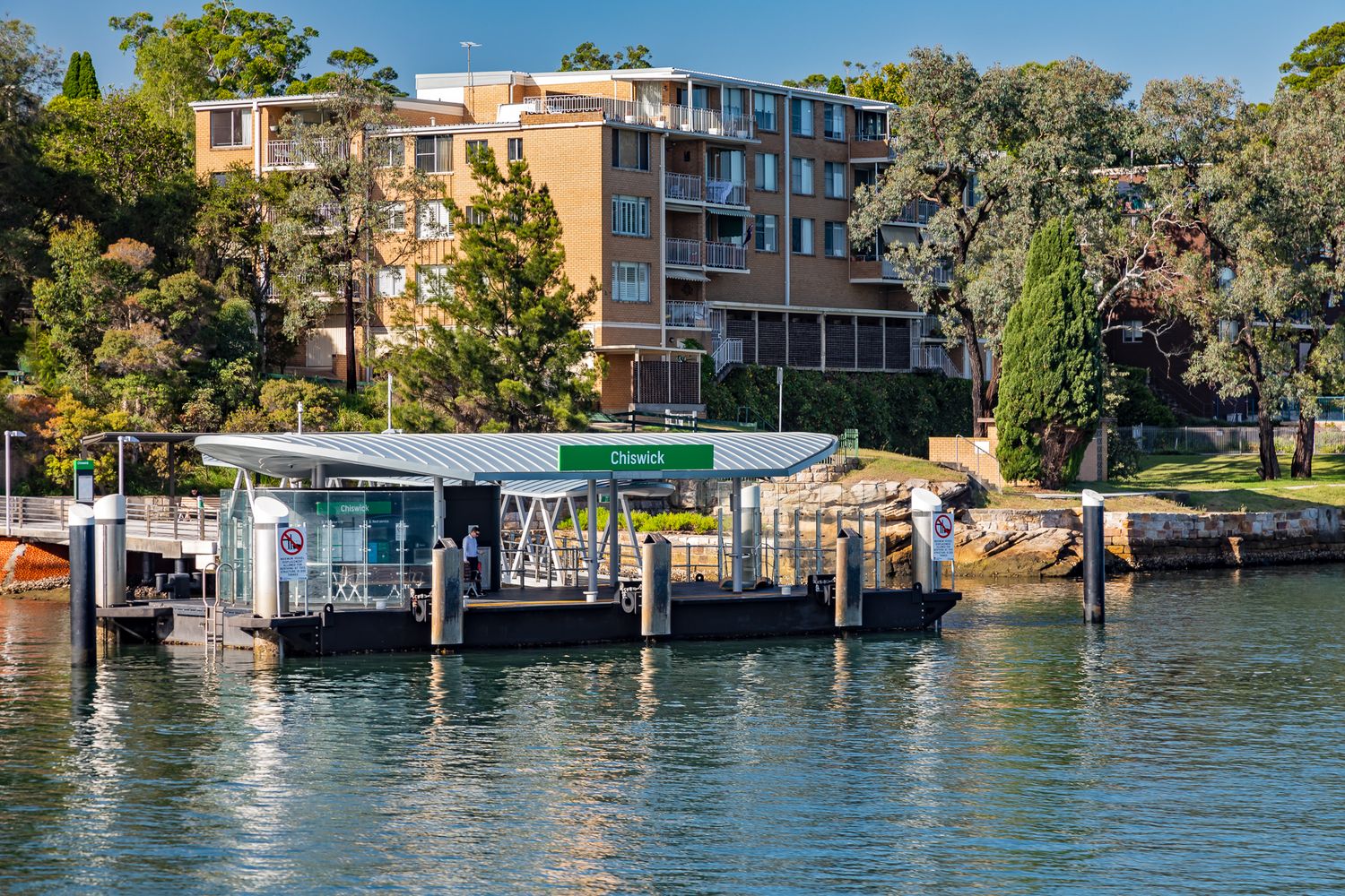 Modern ferry wharf with a green sign on the waterfront, with apartment buildings and leafy trees on the shore behind.