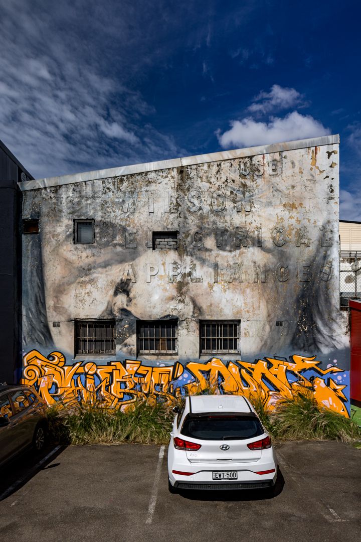 Weathered building wall with a faded painted portrait and orange graffiti lettering above a white hatchback parked in a lot.
