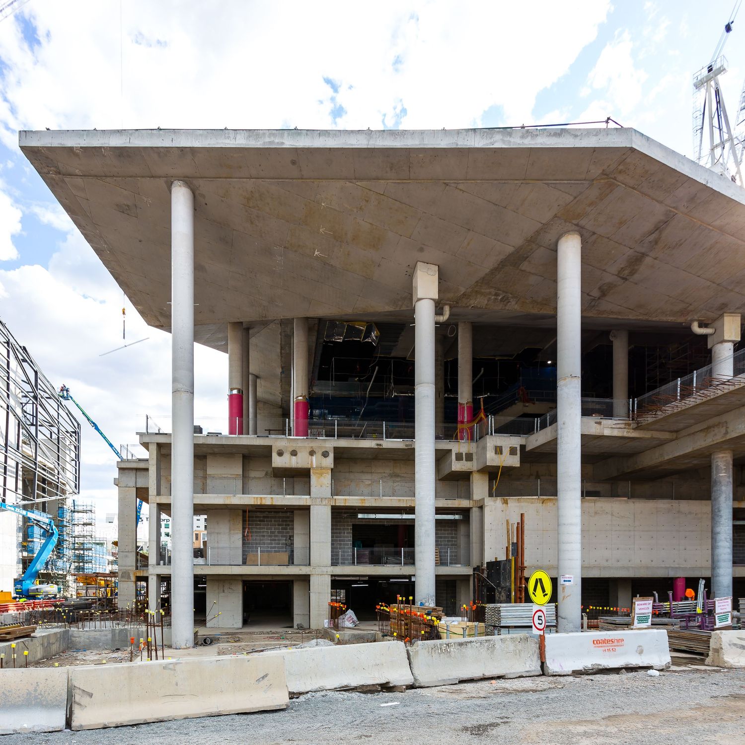 Building under construction with a large concrete canopy supported by tall white columns, barriers and cranes at the site.