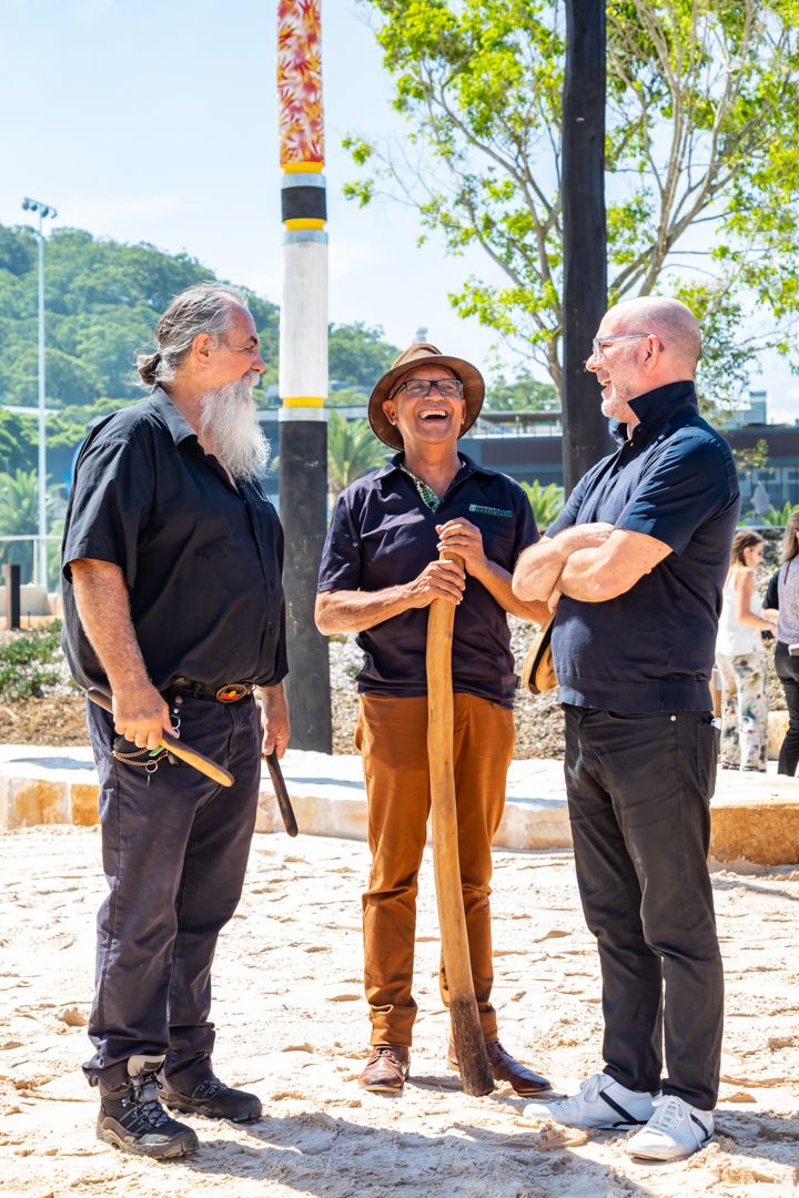 Three men laughing and chatting outdoors on sand, one holding a wooden stick and another a boomerang, with a painted pole behind them.