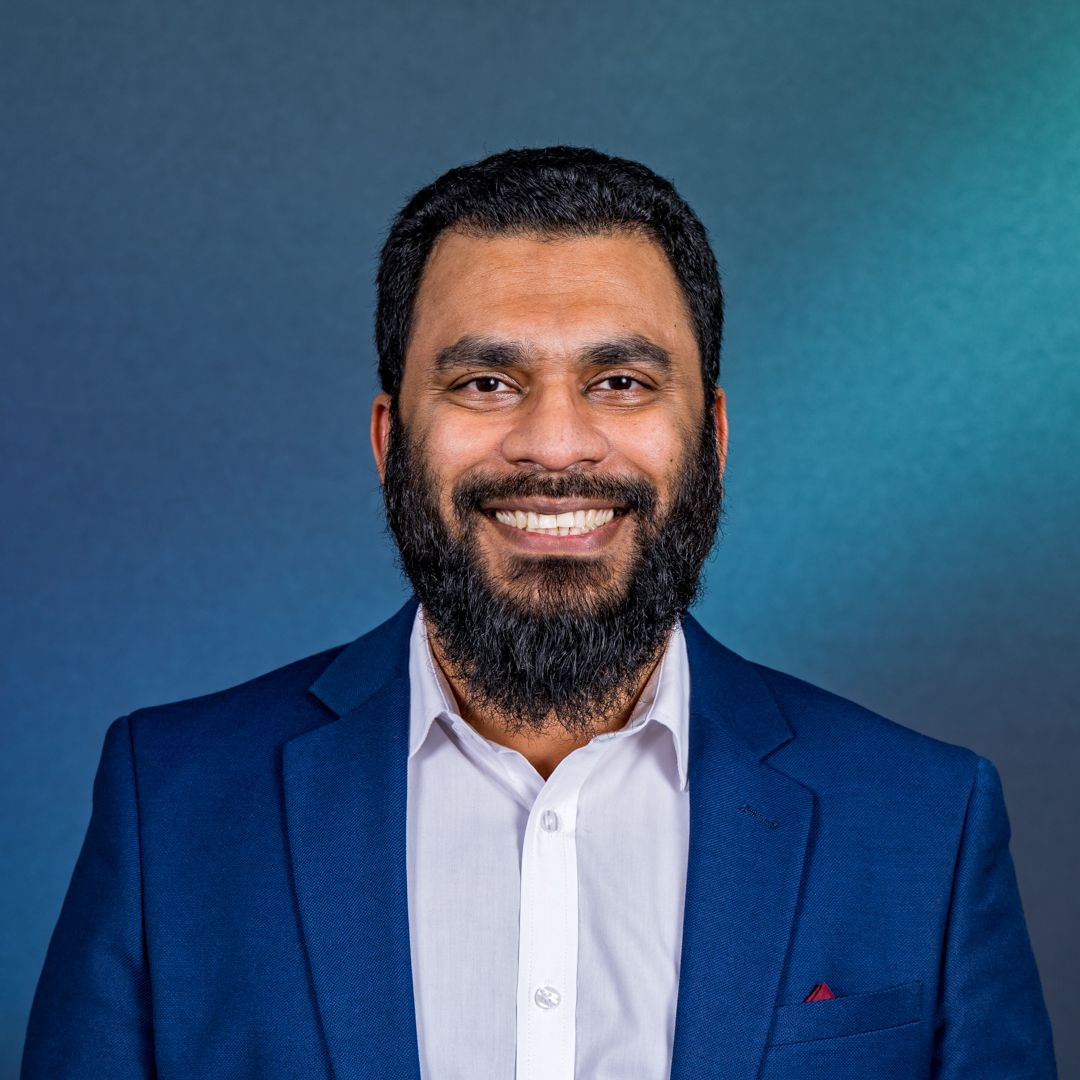 Smiling bearded man in a navy blazer and white collared shirt poses against a blue-teal gradient background in a professional headshot.