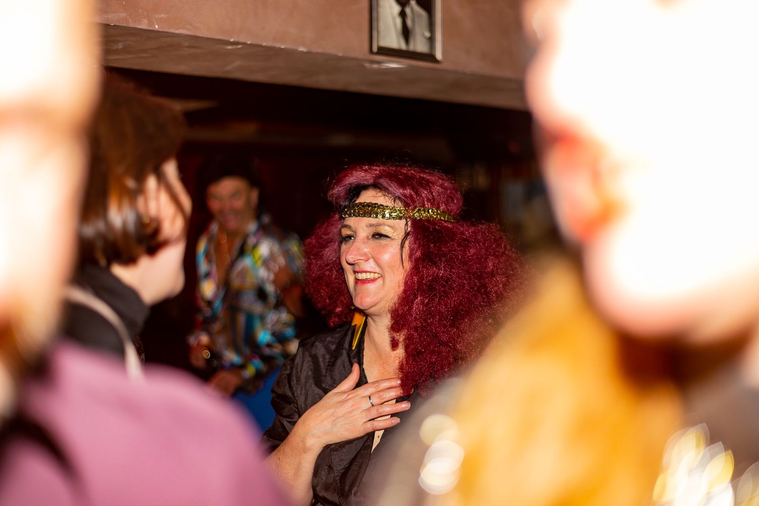 A smiling woman with curly red hair and a gold headband clasps her hands to her chest at a lively party with blurred guests around her.