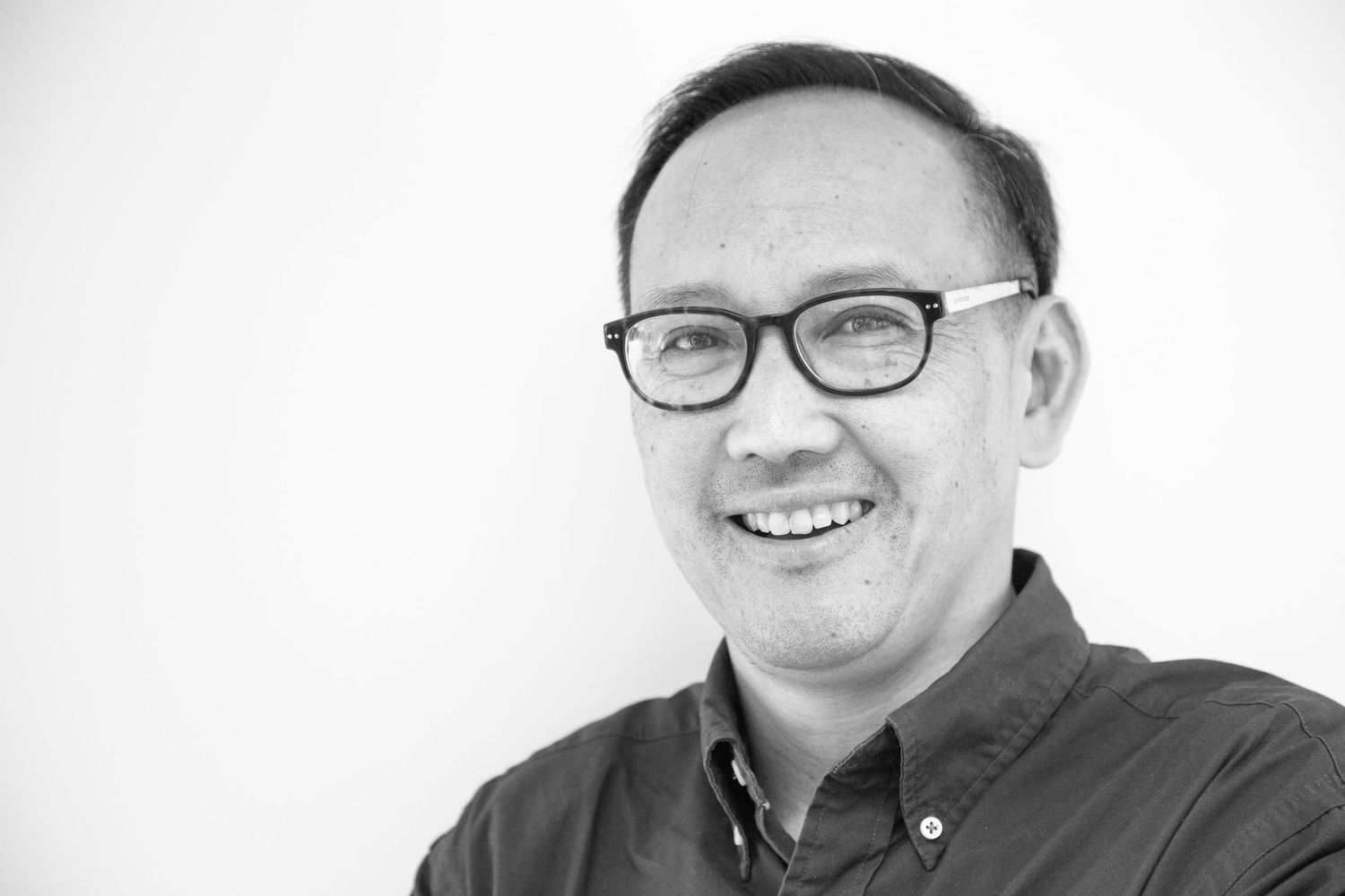 Black and white portrait of a smiling man with dark-rimmed glasses wearing a casual button-up shirt.