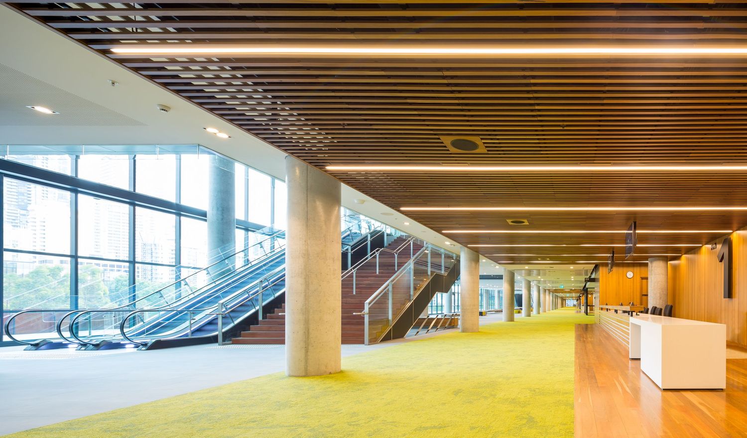 Bright convention centre foyer with timber slat ceiling, linear lighting, escalators, concrete columns and a vibrant green carpet.