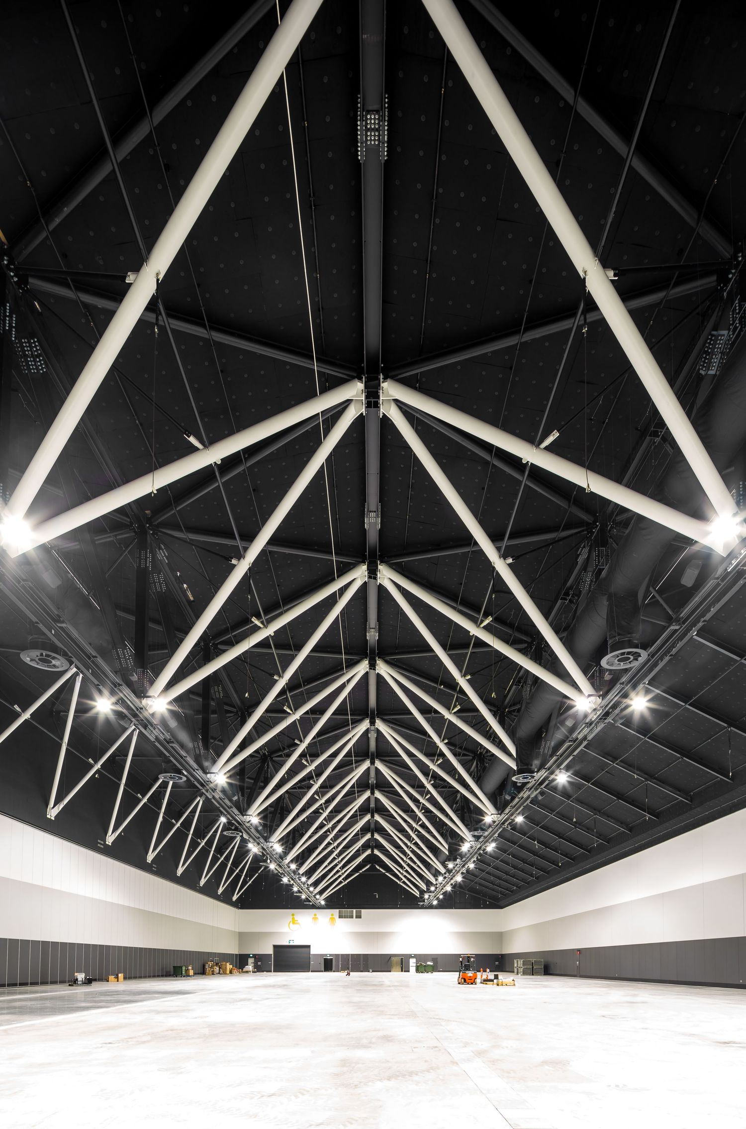 Wide exhibition hall with a dark ceiling supported by white steel trusses and a bright, empty floor below.