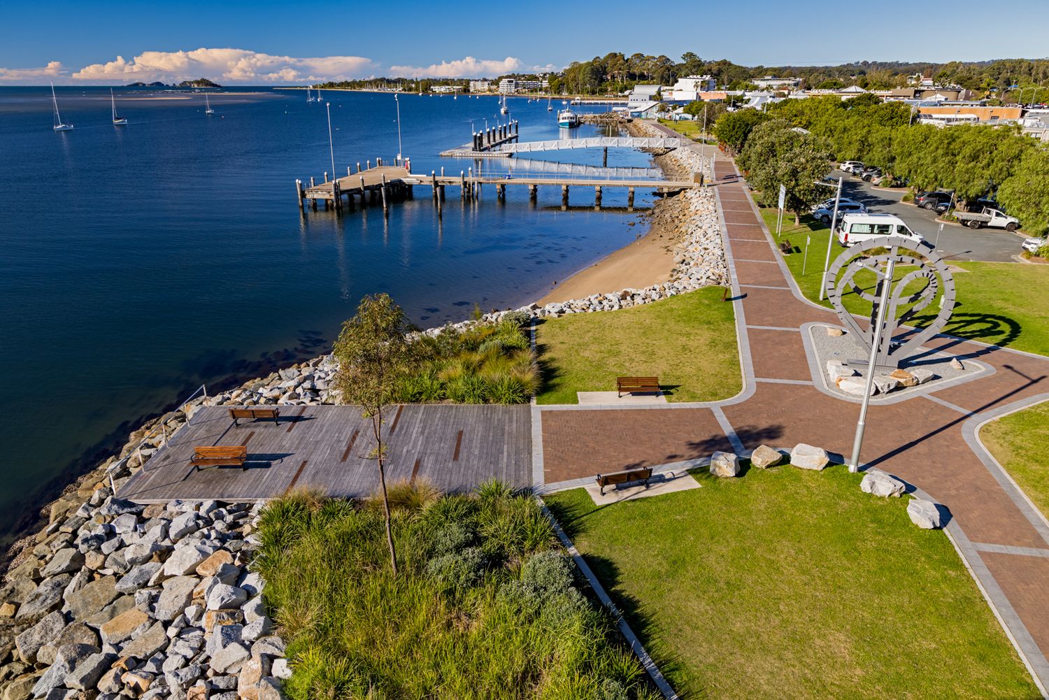 Aerial view of a coastal foreshore park with a timber jetty, rocky shoreline, lawns, walking paths and calm blue bay waters.