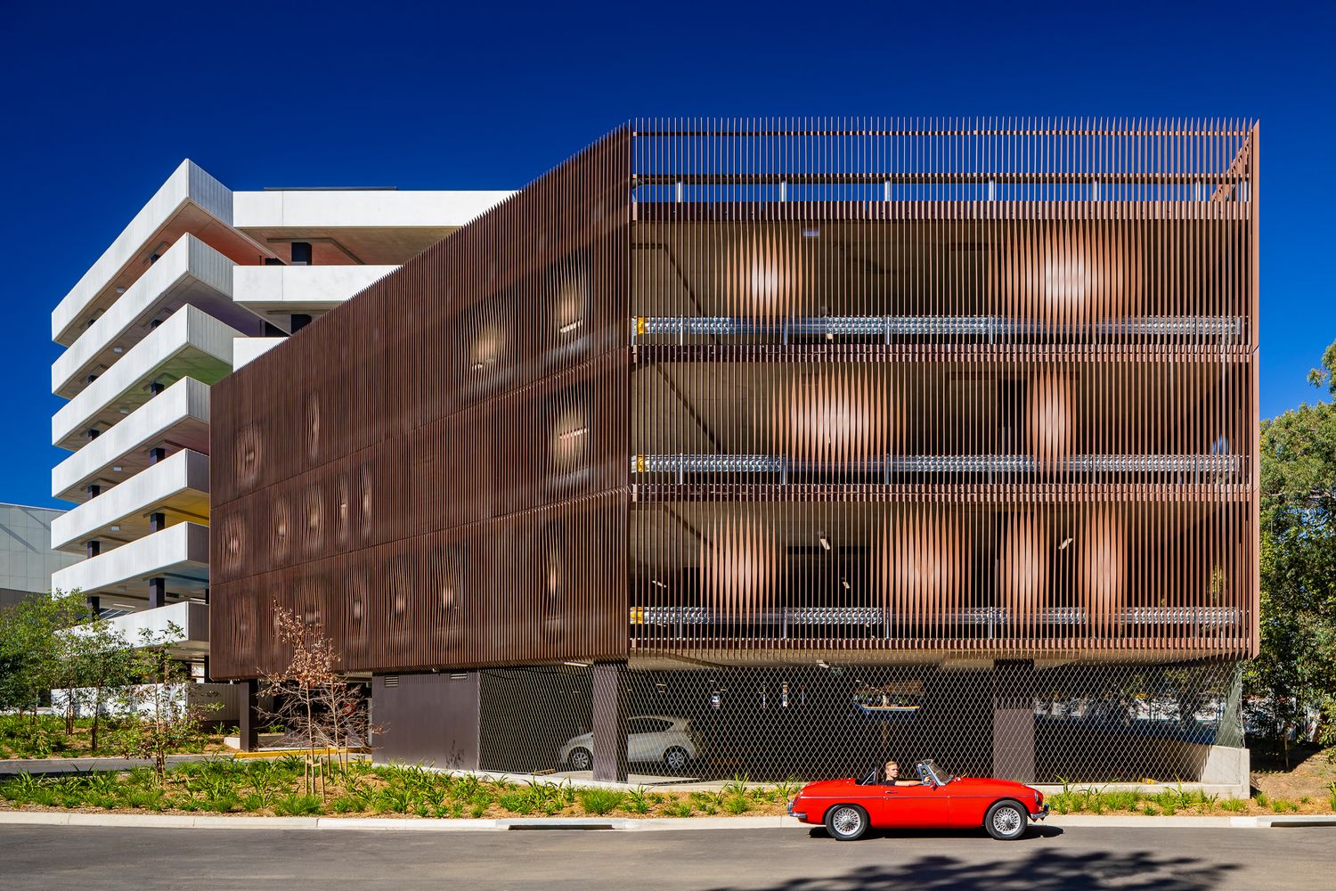Multi-storey car park clad in warm timber-look vertical screens with round openings, with a red vintage convertible parked in front.