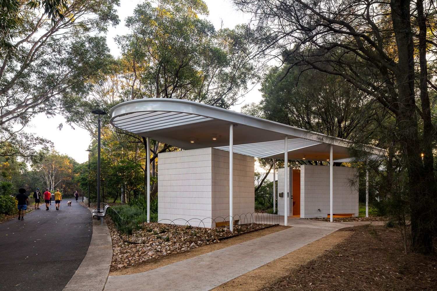 Contemporary park amenities pavilion with a curved white roof and tiled walls beside a walking path and trees.