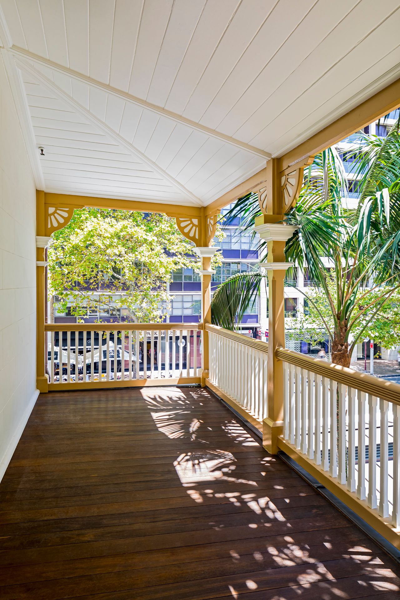 Corner of a restored heritage verandah with ornate fretwork, white balustrade and dark timber floorboards dappled in sunlight.
