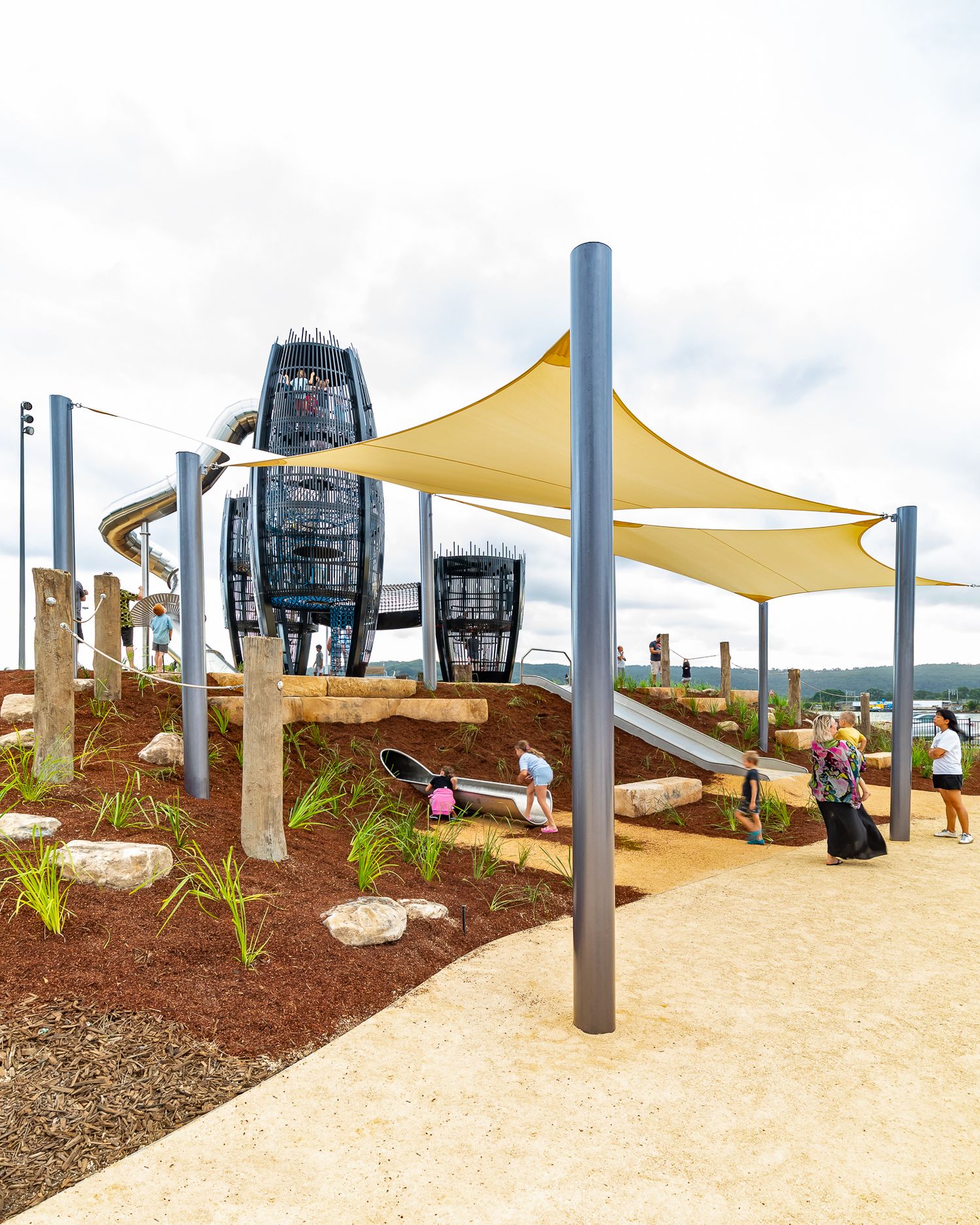 Playground with sail shade canopies, landscaped garden beds, timber posts and tall climbing towers as families walk the sandy path.